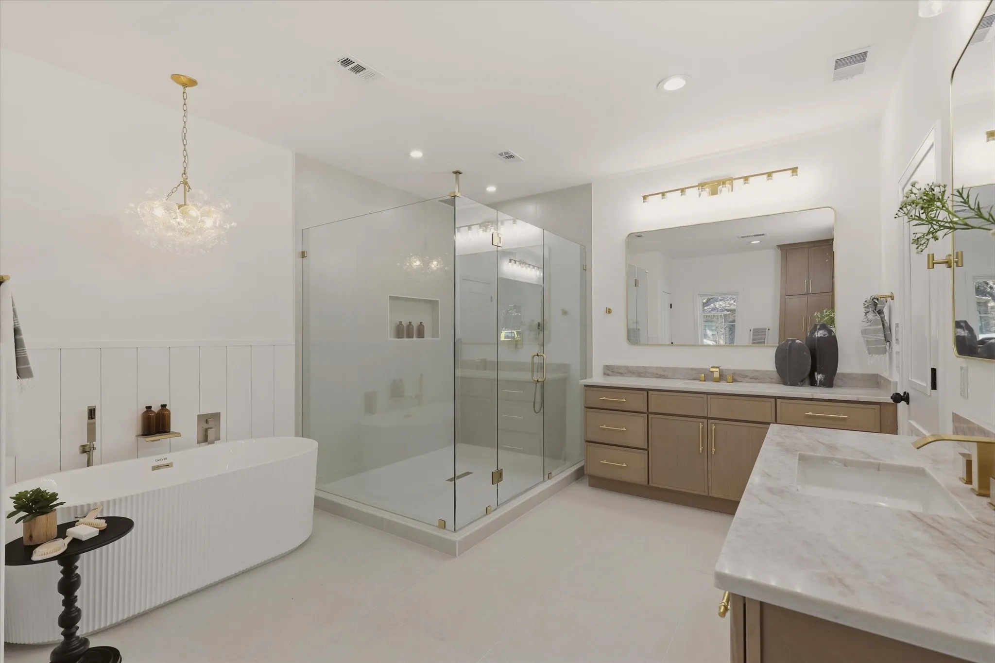 Freestanding soaking tub with vertical fluting, a spacious glass-enclosed shower, dual vanity with undermount sinks, stone countertops, and gold-toned hardware