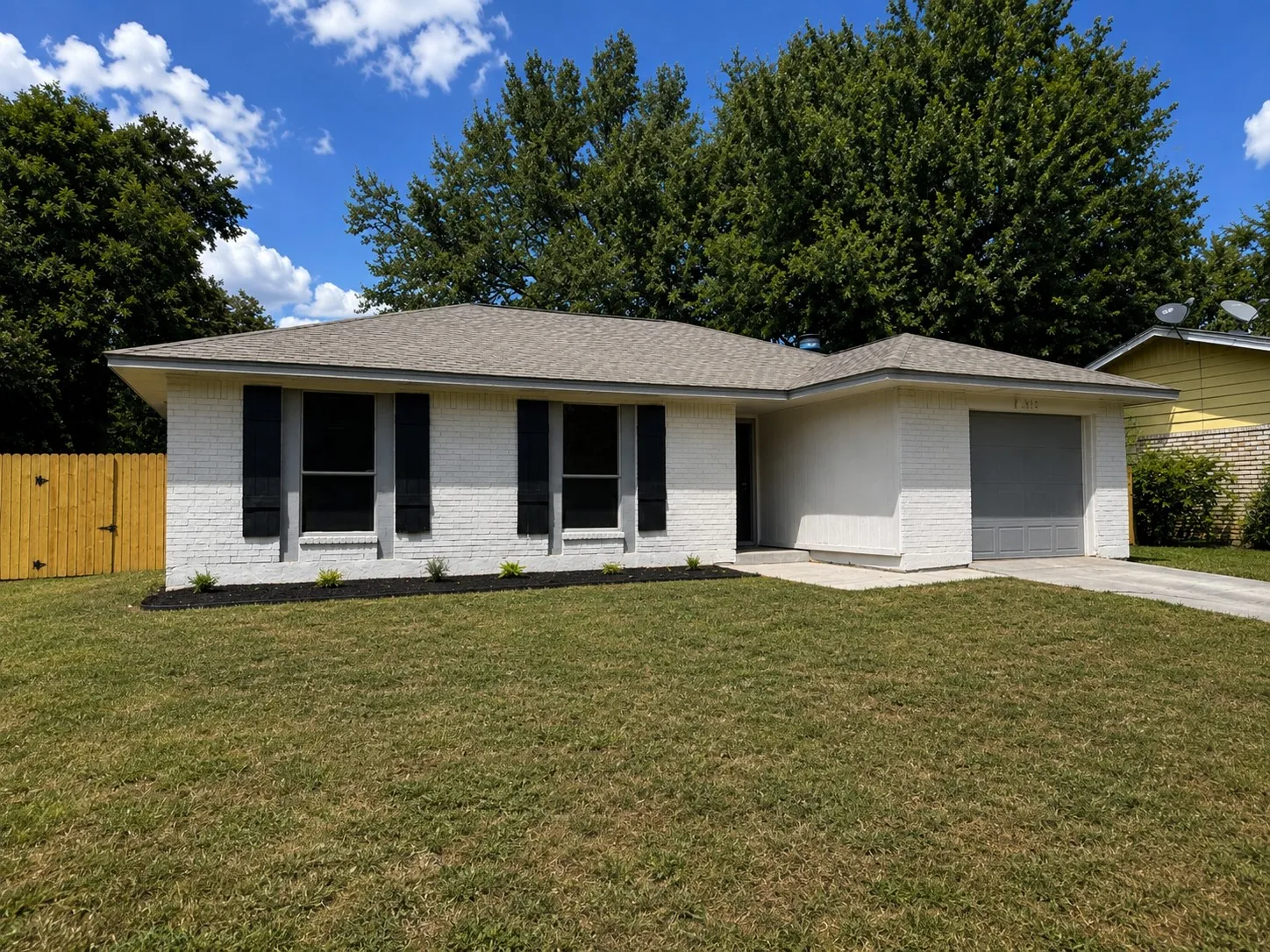 White brick exterior with black window shutters, attached garage, front landscaping, and a wood privacy​​‌​​​​‌​​‌‌​‌‌​​​‌‌​‌​‌​‌​​​‌​​ fence