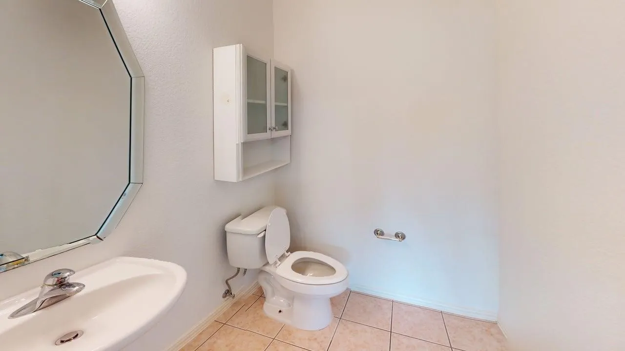 Pedestal sink with chrome faucet, octagonal mirror, wall-mounted storage cabinet with glass-front doors, toilet, and tiled flooring