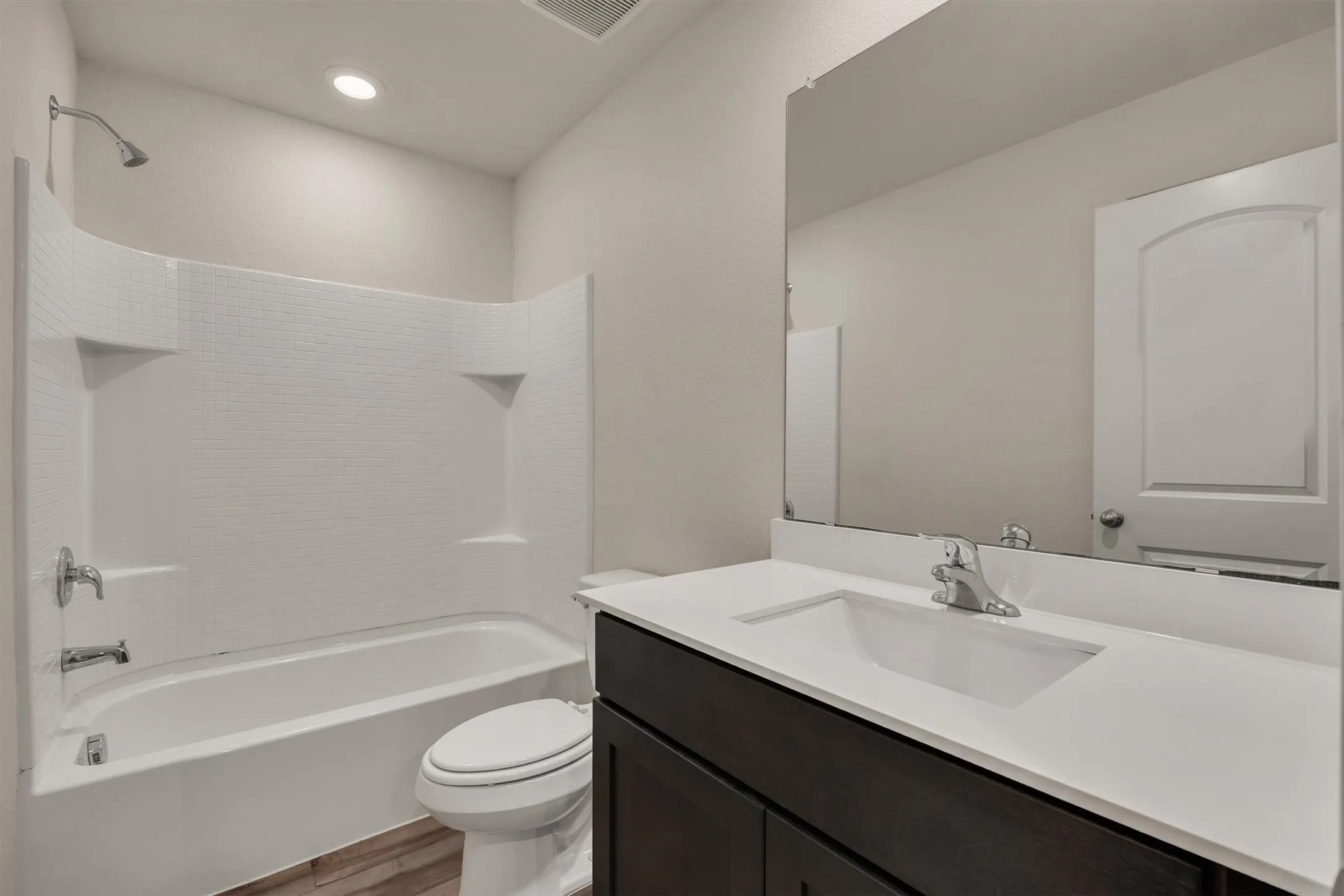 White bathtub with integrated shower, dark wood-finish vanity with white countertop, and expansive wall-mounted mirror