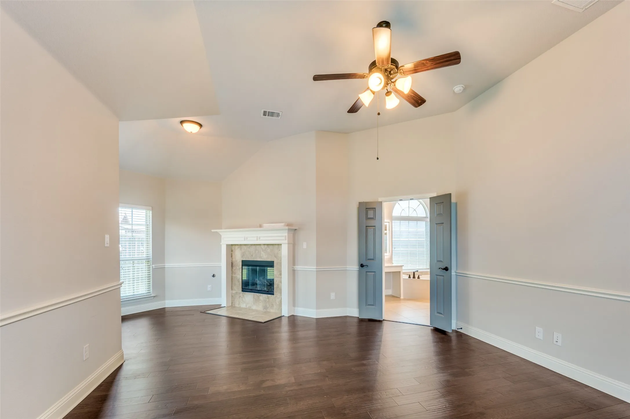 Dark wood-finish flooring, vaulted ceiling, built-in fireplace with tile surround, and a ceiling fan with integrated lighting