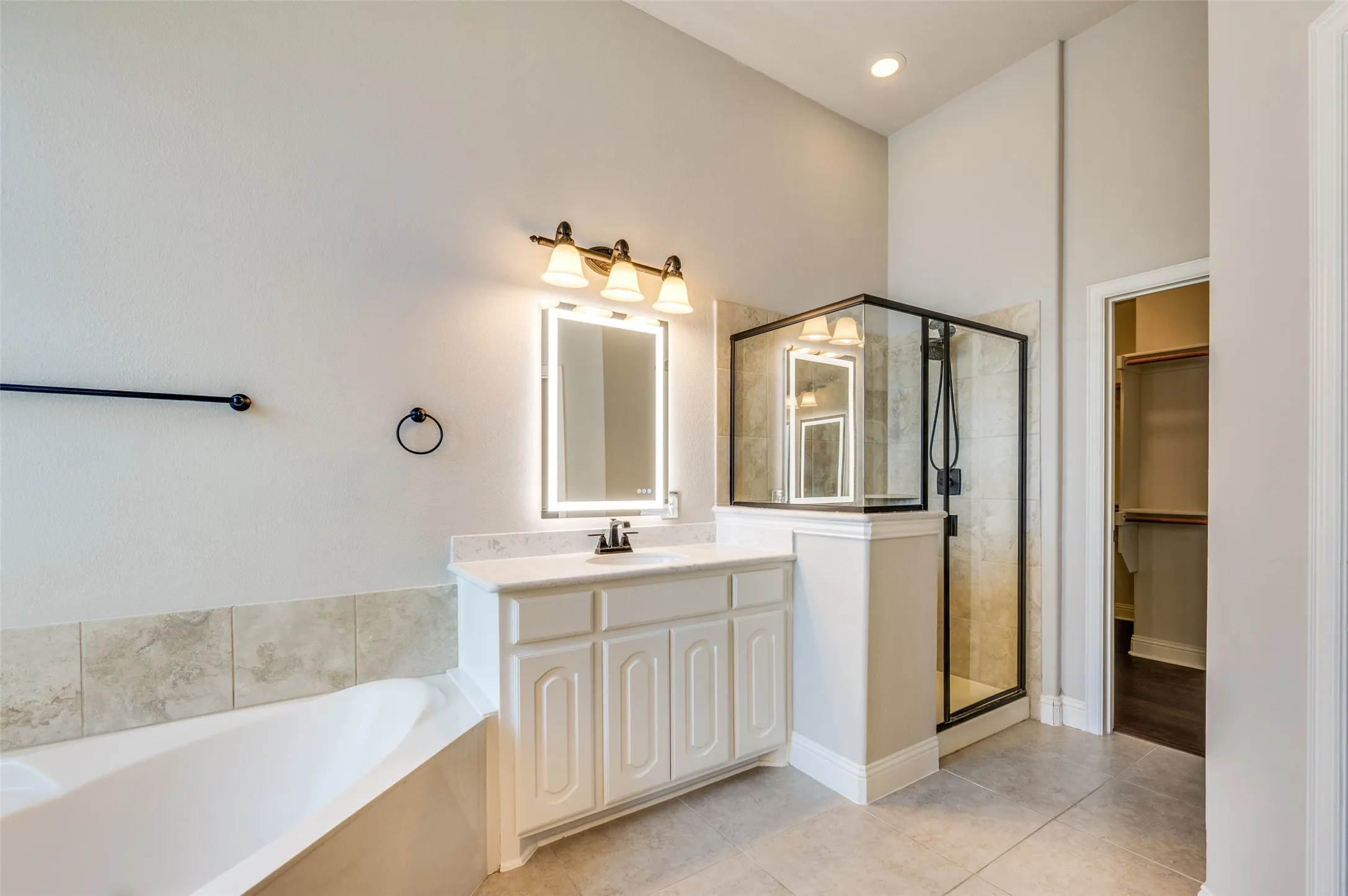 White vanity with a single basin sink and illuminated mirror, large soaking tub with tiled surround, and walk-in shower with black-framed glass enclosure