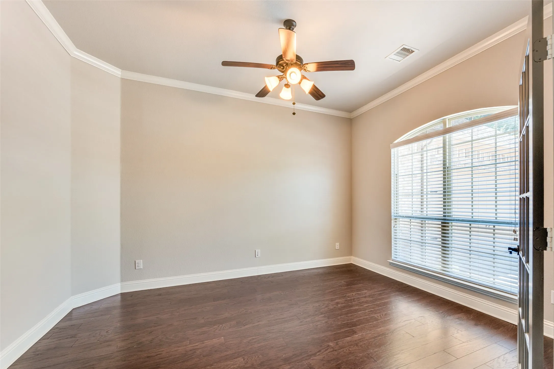 Hardwood-finish flooring throughout, ceiling fan with integrated lighting, arched window with blinds, crown molding, and a solid-core door with decorative ironwork