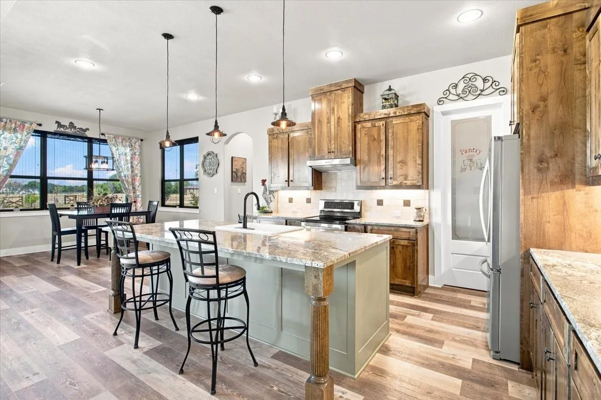 Open-concept kitchen with wood-finish flooring, a central island featuring a dual-basin sink and pendant lighting, wood cabinetry, and a pantry with a frosted glass door