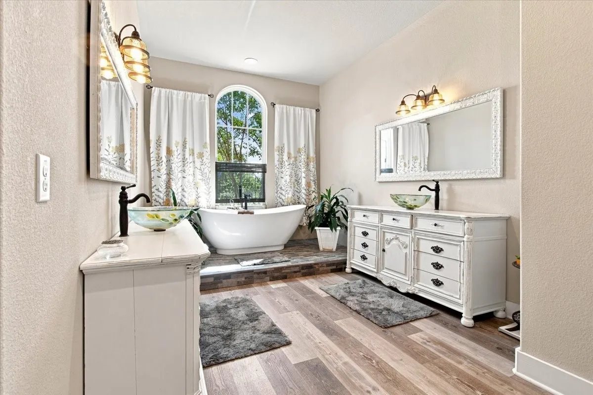 Wood-finish flooring with a freestanding soaking tub, dual vanity consoles, and an arched window