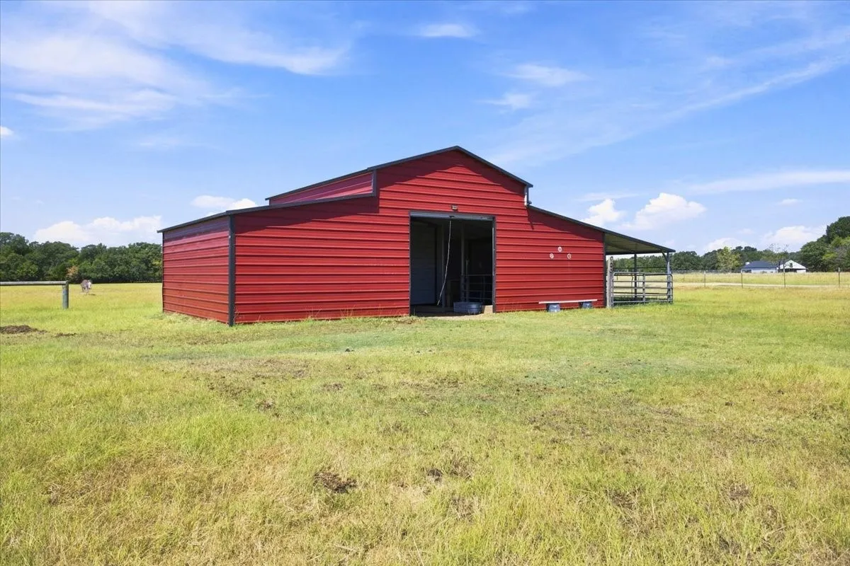 Red metal barn with an open entryway, covered side area, and surrounding grassy expanse