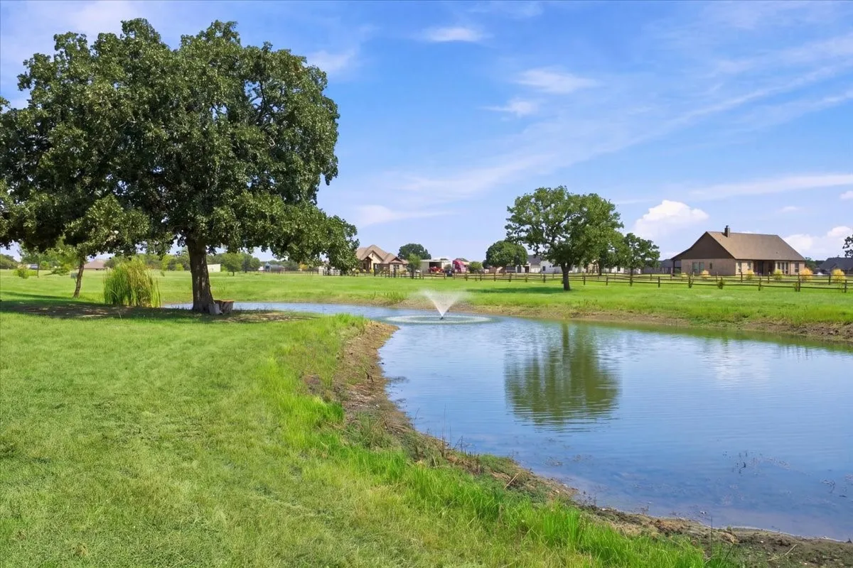 Pond with a fountain feature, expansive grassy areas, mature trees, and a distant property with a pitched roof