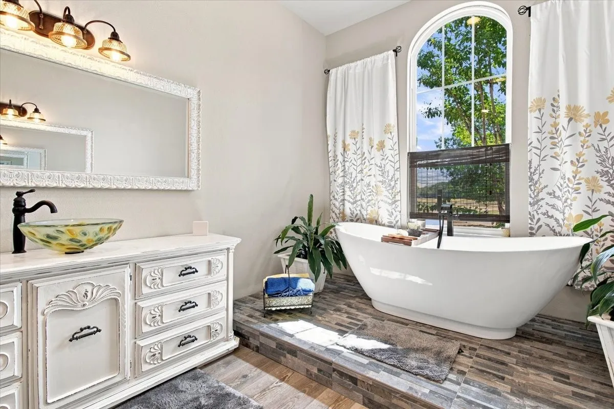 Freestanding soaking tub, arched window, wood-finish flooring, vanity with vessel sink, and an ornate framed mirror