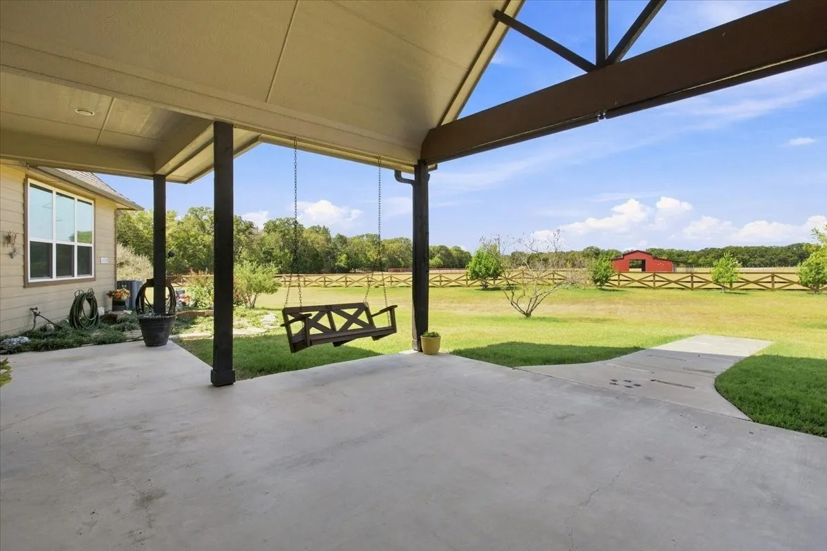 Expansive covered patio featuring a porch swing, vaulted ceiling, and concrete flooring, overlooking a green space with a red barn