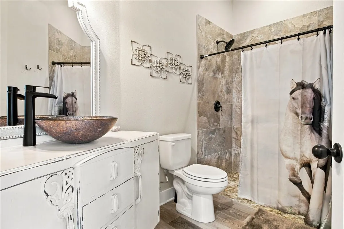 Distressed white vanity with decorative elements, vessel sink with black faucet, tiled shower surround, white toilet, and wood-finish flooring