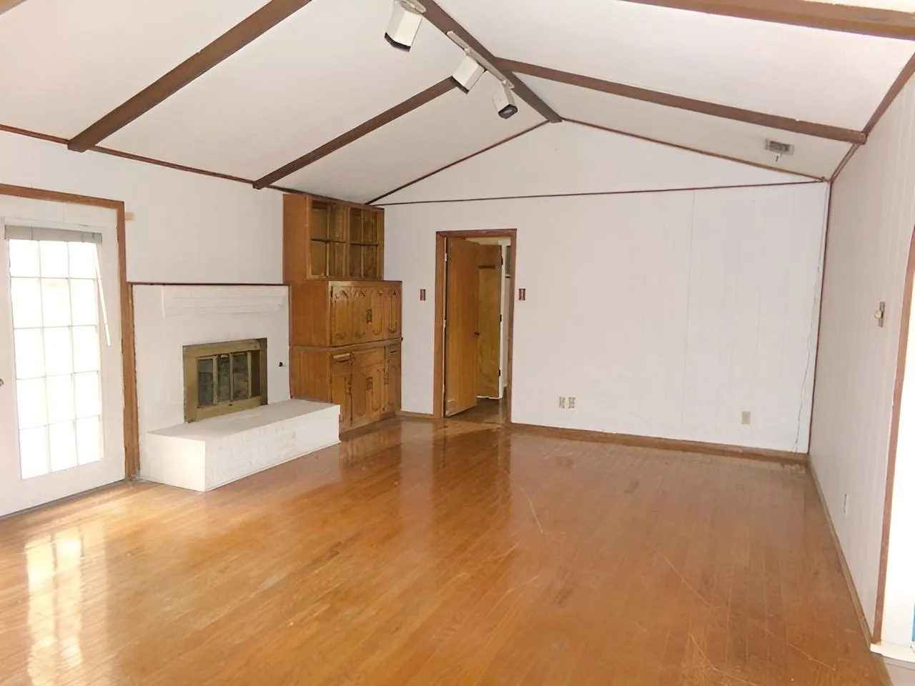 Vaulted ceiling with exposed beams, wood-finish flooring, white brick fireplace with a brass firebox, built-in wood cabinetry, and a door with glass block inserts