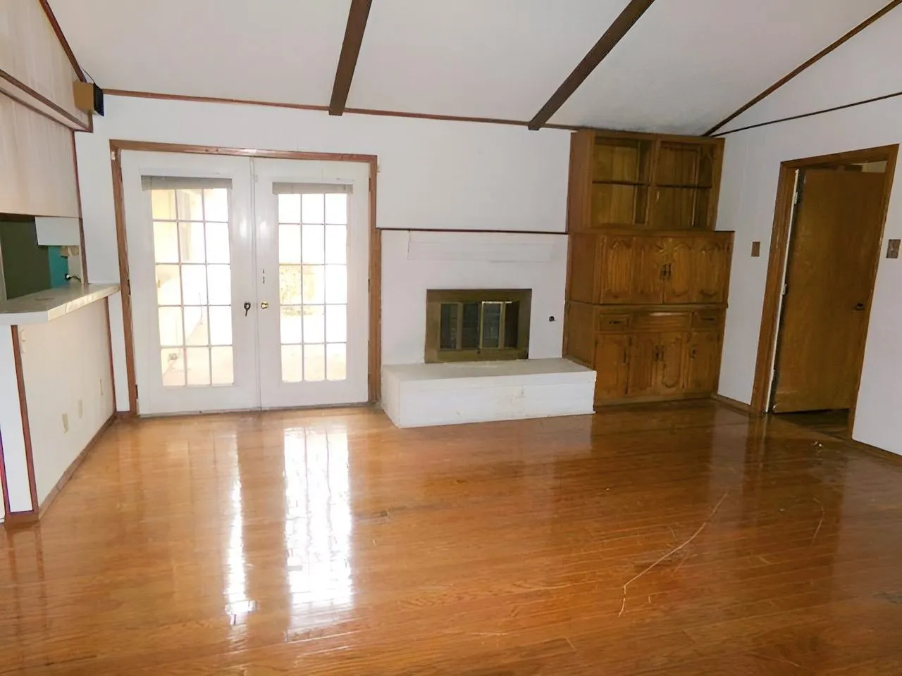 Vaulted ceiling with exposed beams, wood-finish flooring, white fireplace with hearth, built-in wooden cabinetry, and French doors