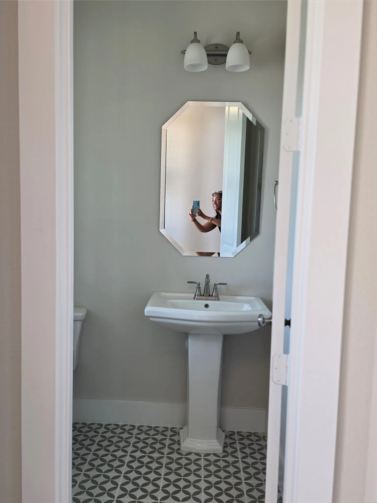 Pedestal sink with chrome faucet, octagonal mirror, two-light vanity fixture, and patterned floor tile