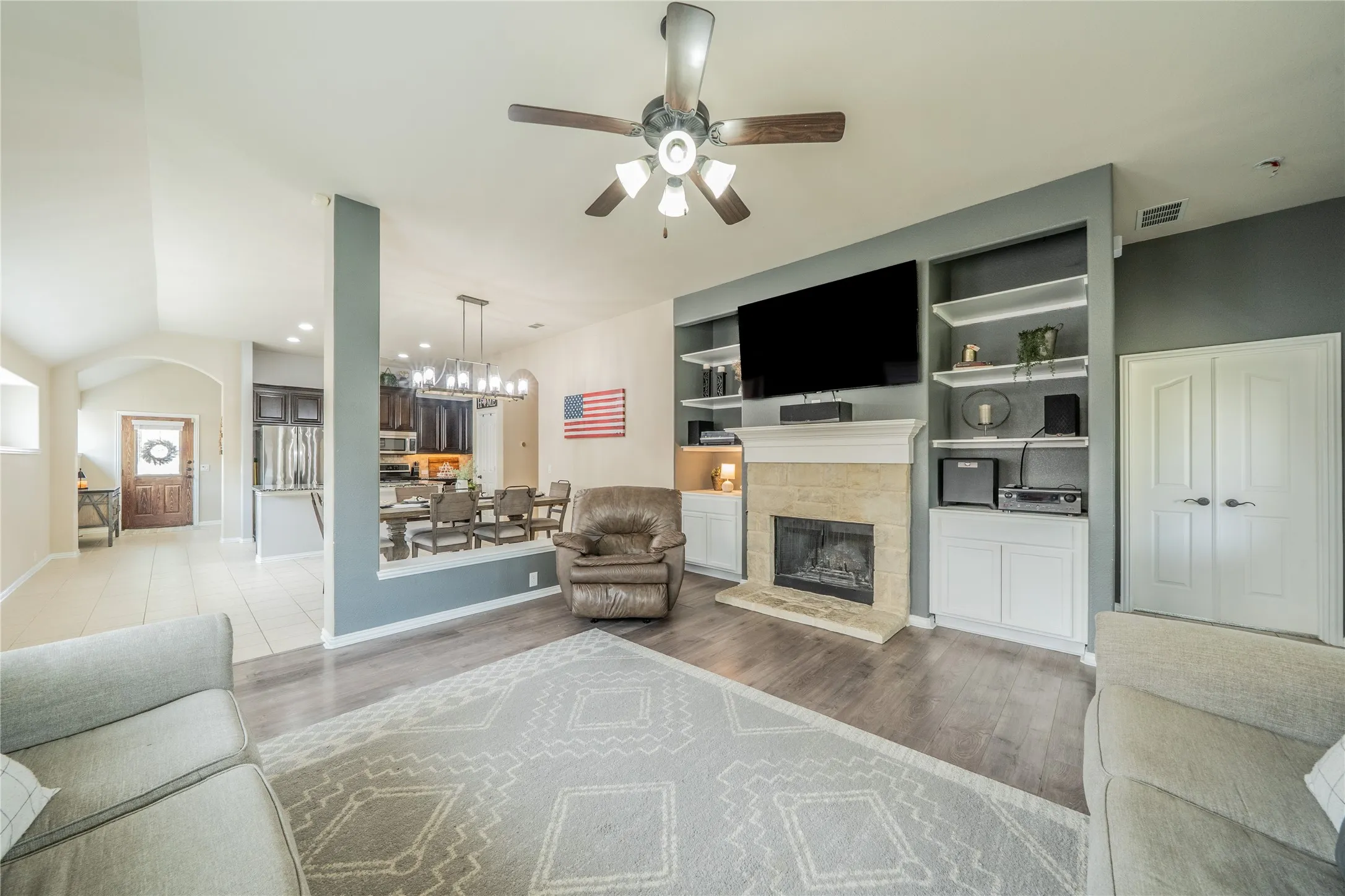 Living area featuring wood-finish flooring, a ceiling fan, and a stone-clad fireplace with built-in shelving