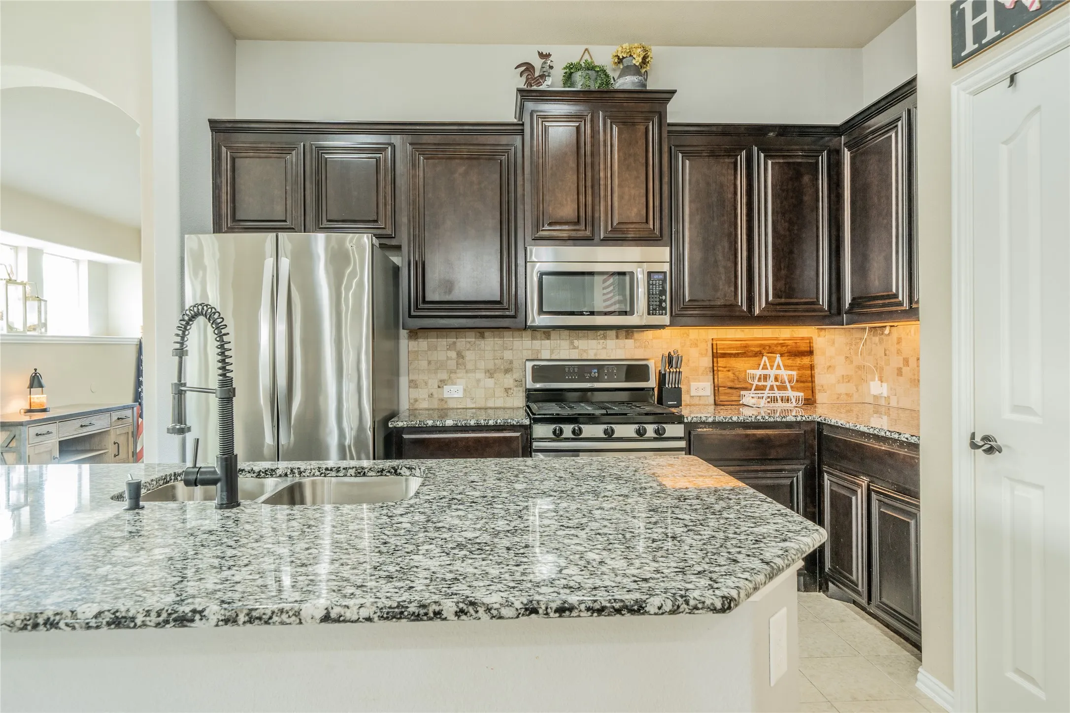 Spacious kitchen featuring an island with a stainless steel sink and a gooseneck faucet, granite countertops, dark wood cabinetry, stainless steel appliances, and a tiled backsplash