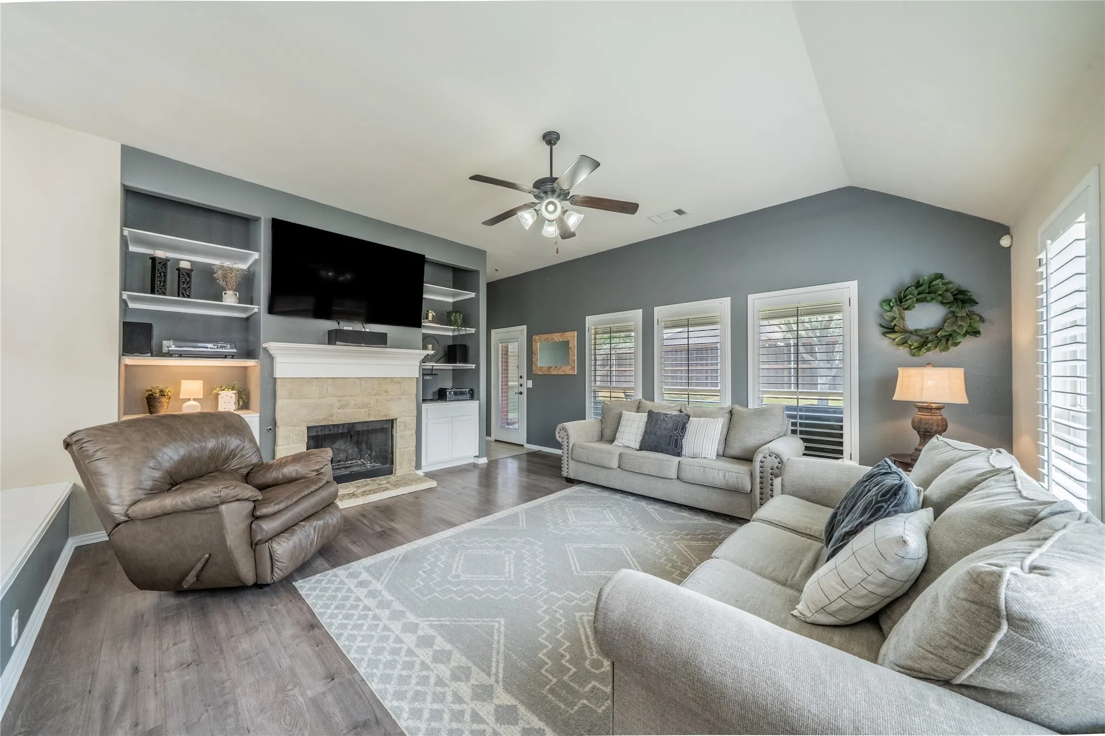 Vaulted ceiling living area featuring wood-finish flooring, a stone-surround fireplace with mantel, built-in shelving, and multiple windows with plantation shutters