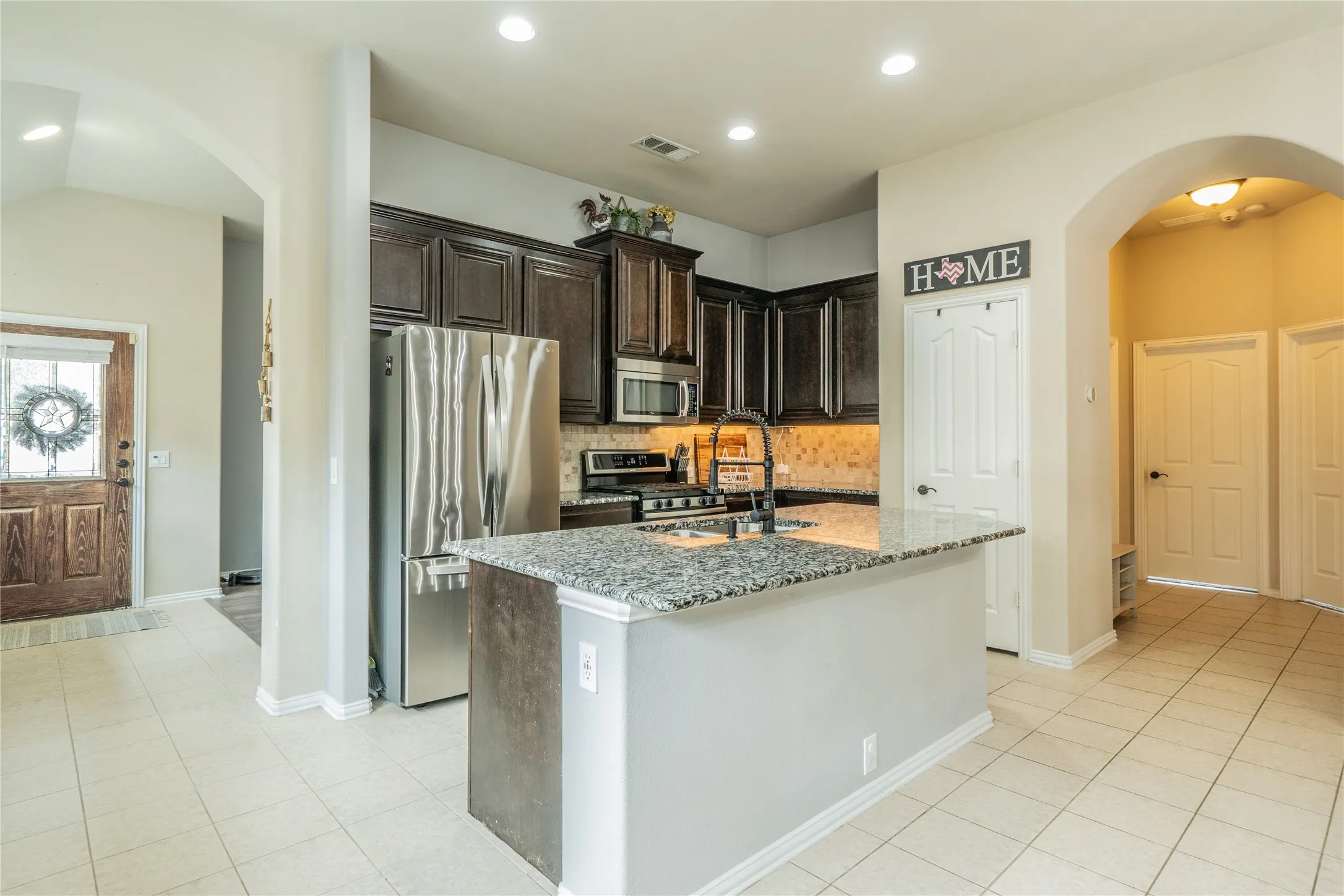 Kitchen featuring dark wood cabinetry, stainless steel appliances, a granite-topped island with an integrated sink, and tile flooring
