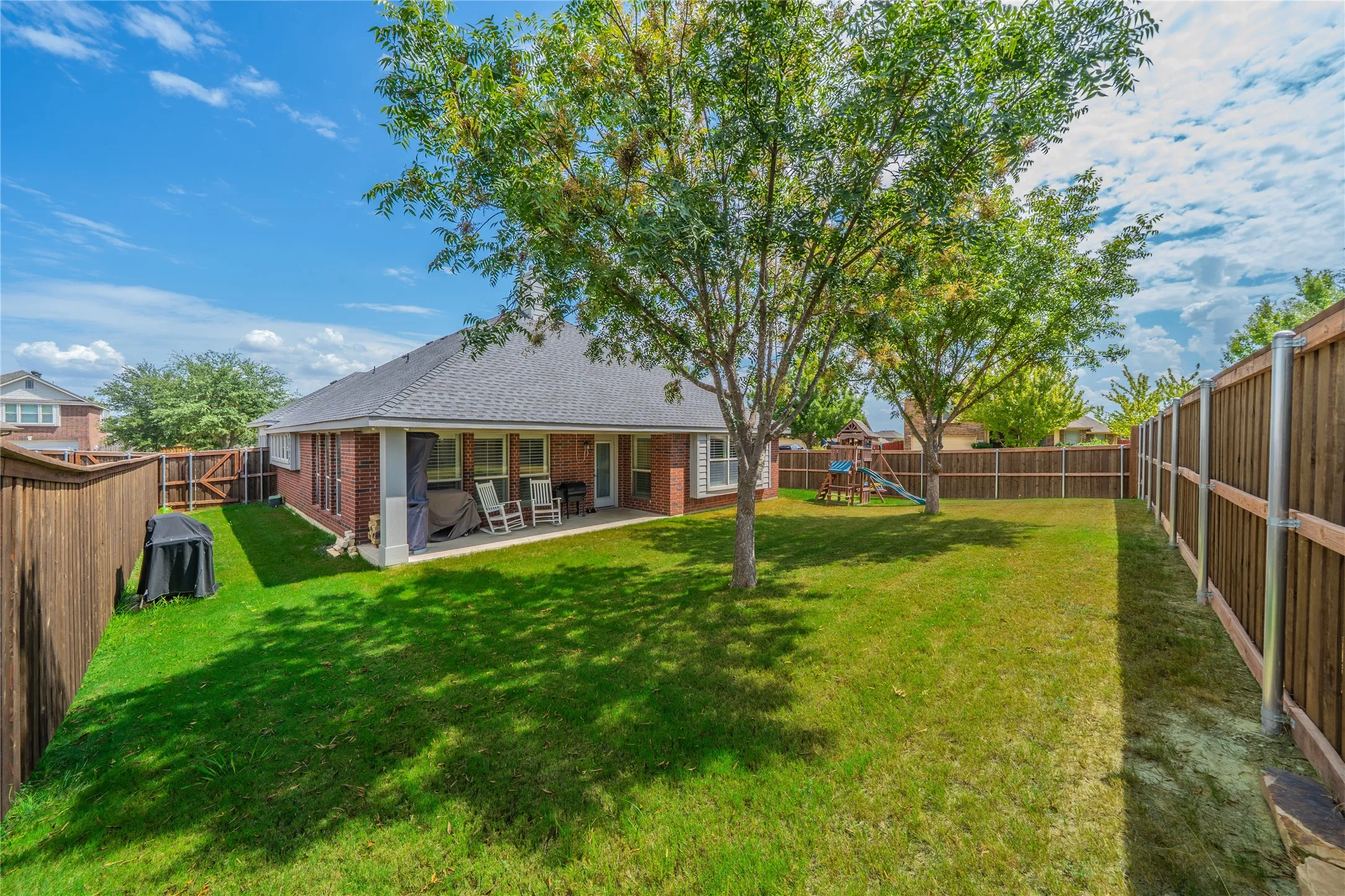 Expansive fenced backyard featuring a covered patio, brick exterior, and mature trees