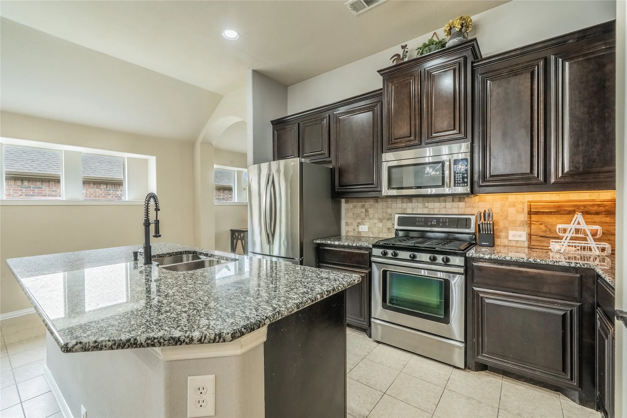 Kitchen featuring dark wood cabinetry, stainless steel appliances, granite countertops, a tiled backsplash, and an island with a sink