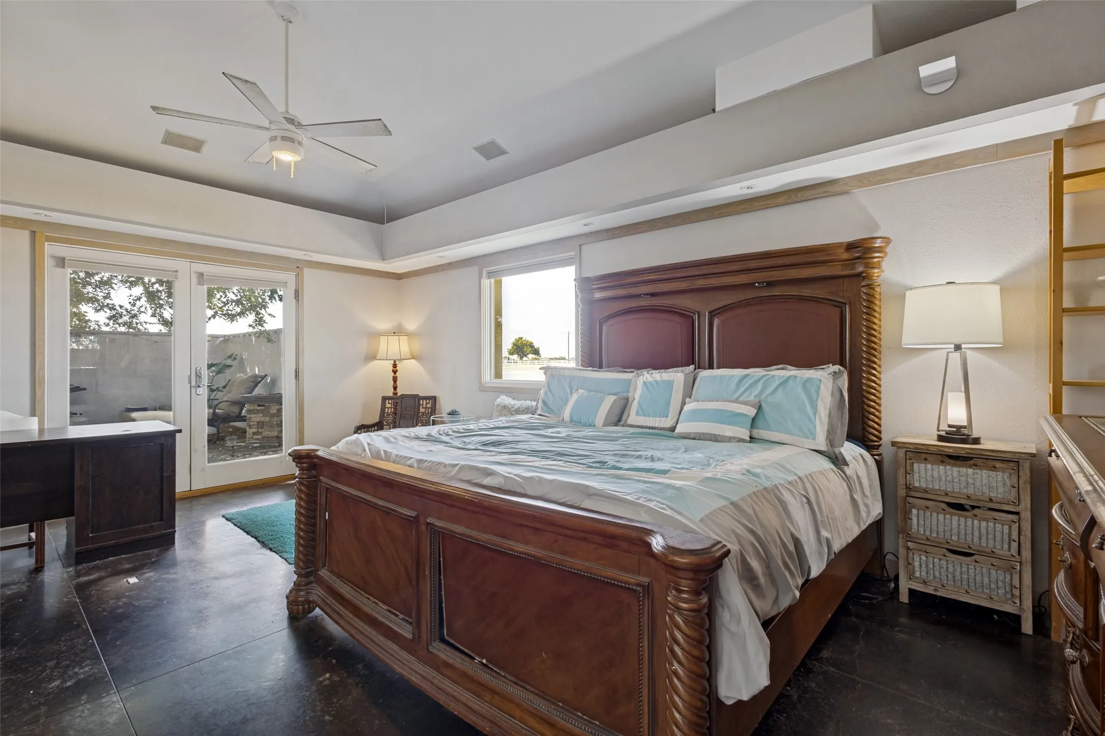 The primary bedroom has abundant space for furniture along with room for a sitting area or desk.  Stained concrete floors are below and a ceiling fan is above.  French doors open to the the patio and additional light comes from a window.