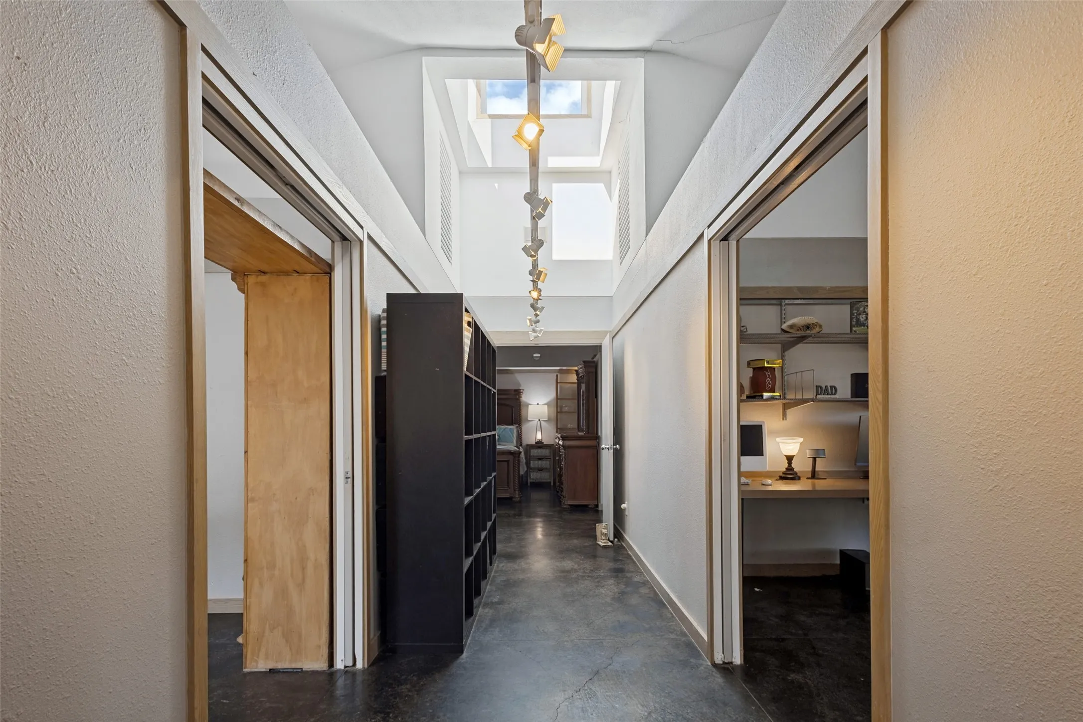 This hallway leads past a bedroom and the office to the primary suite.  The track lighting above and the towering open space containing the skylight all provide striking architectural interest.