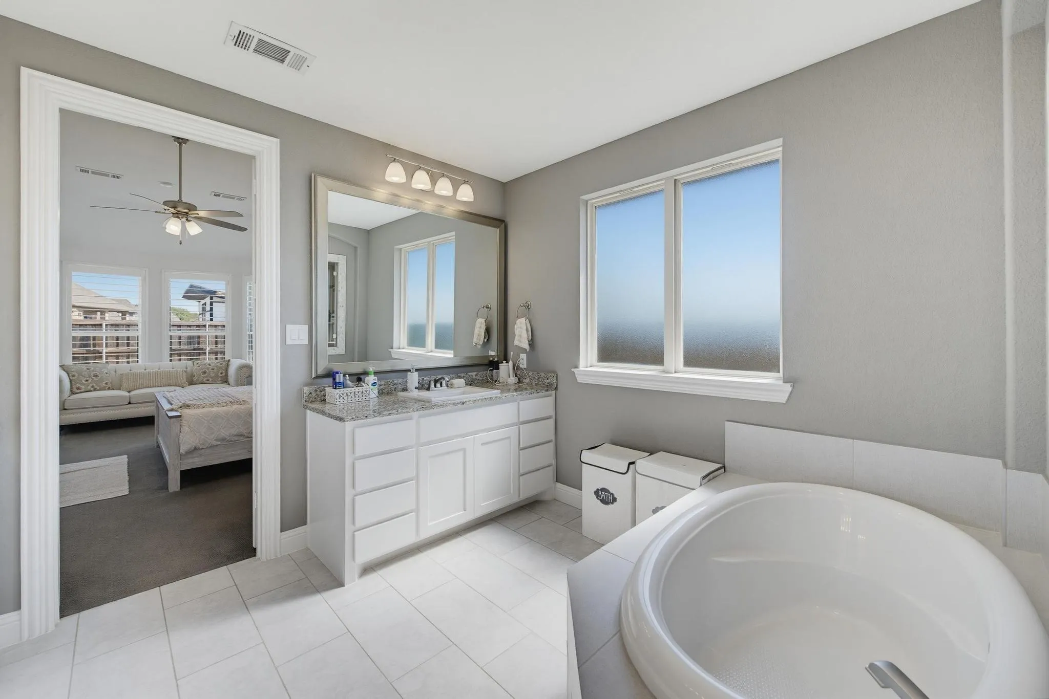 White tile flooring, a built-in soaking tub, a vanity with a stone countertop, an integrated sink, and an expansive wall mirror
