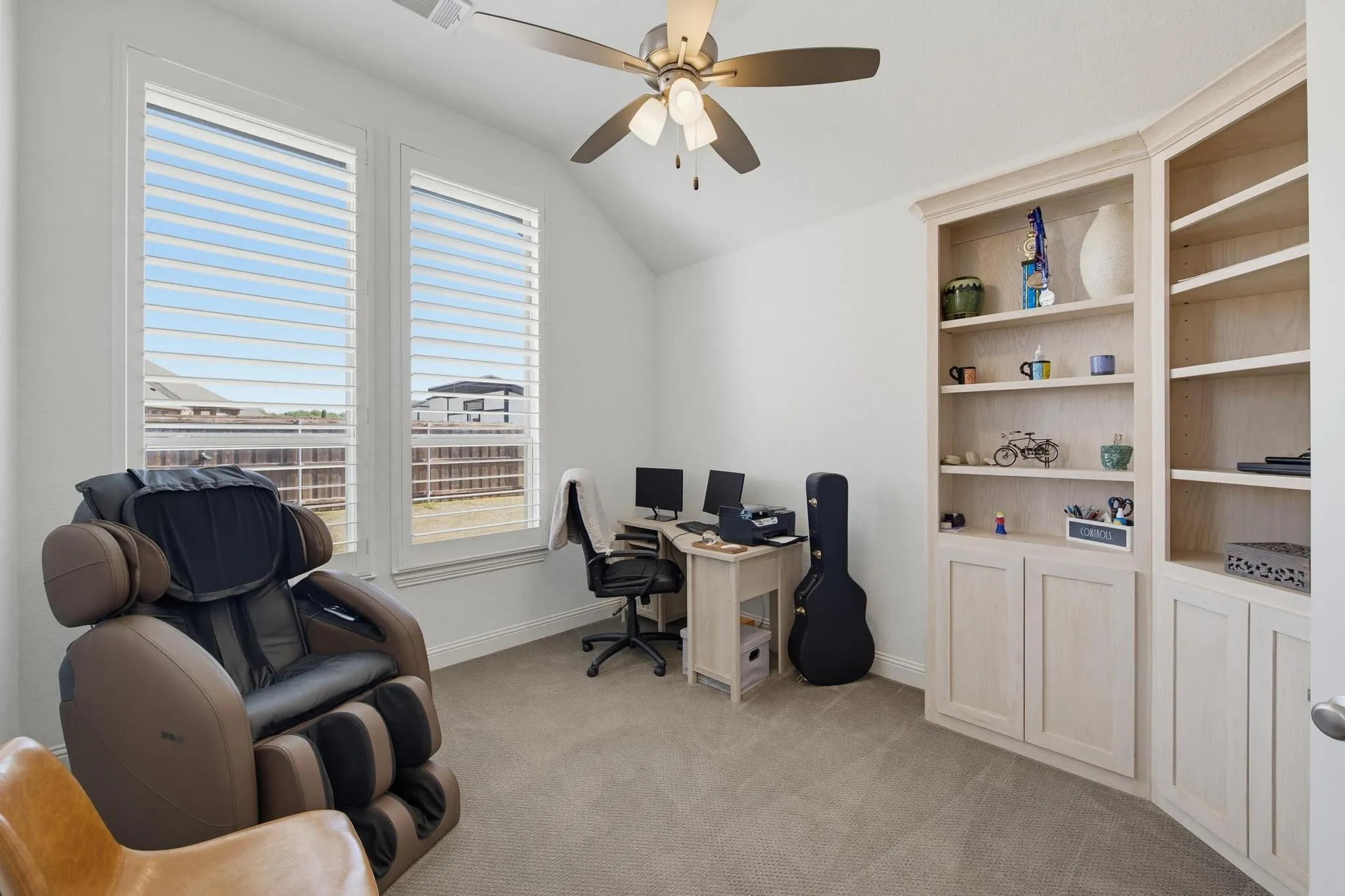 Vaulted ceiling with a ceiling fan, dual windows featuring louvered shutters, light-toned carpet flooring, and custom built-in shelving with cabinetry
