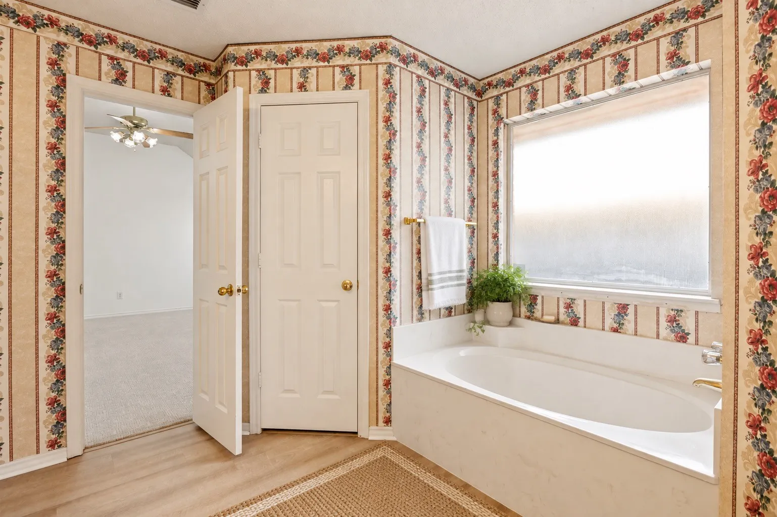 Soaking tub with a large window, brass fixtures, and patterned wallpaper