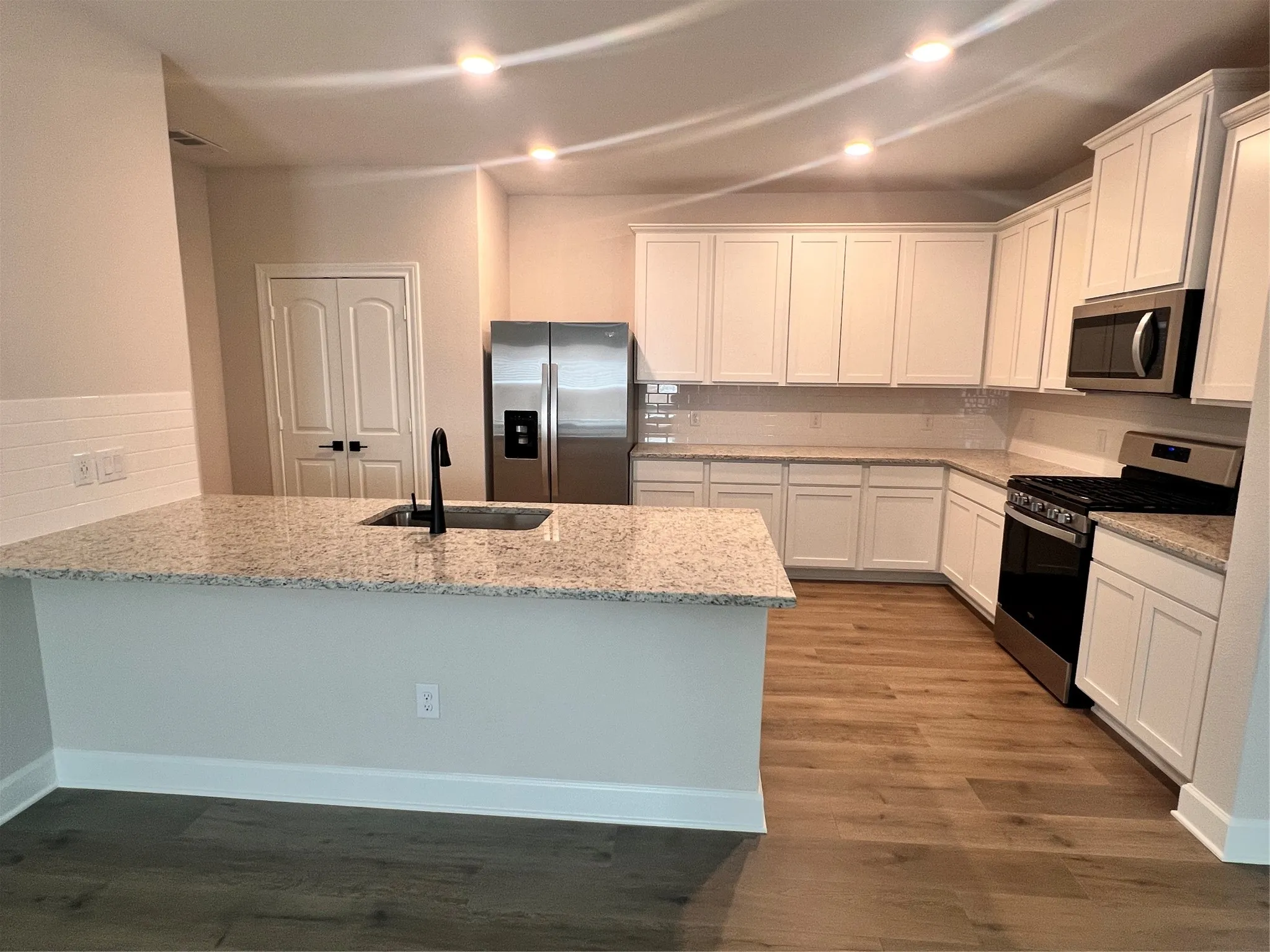 Kitchen featuring an island with an undermount sink, light-toned countertops, white shaker cabinetry, stainless steel appliances, and wood-finish flooring