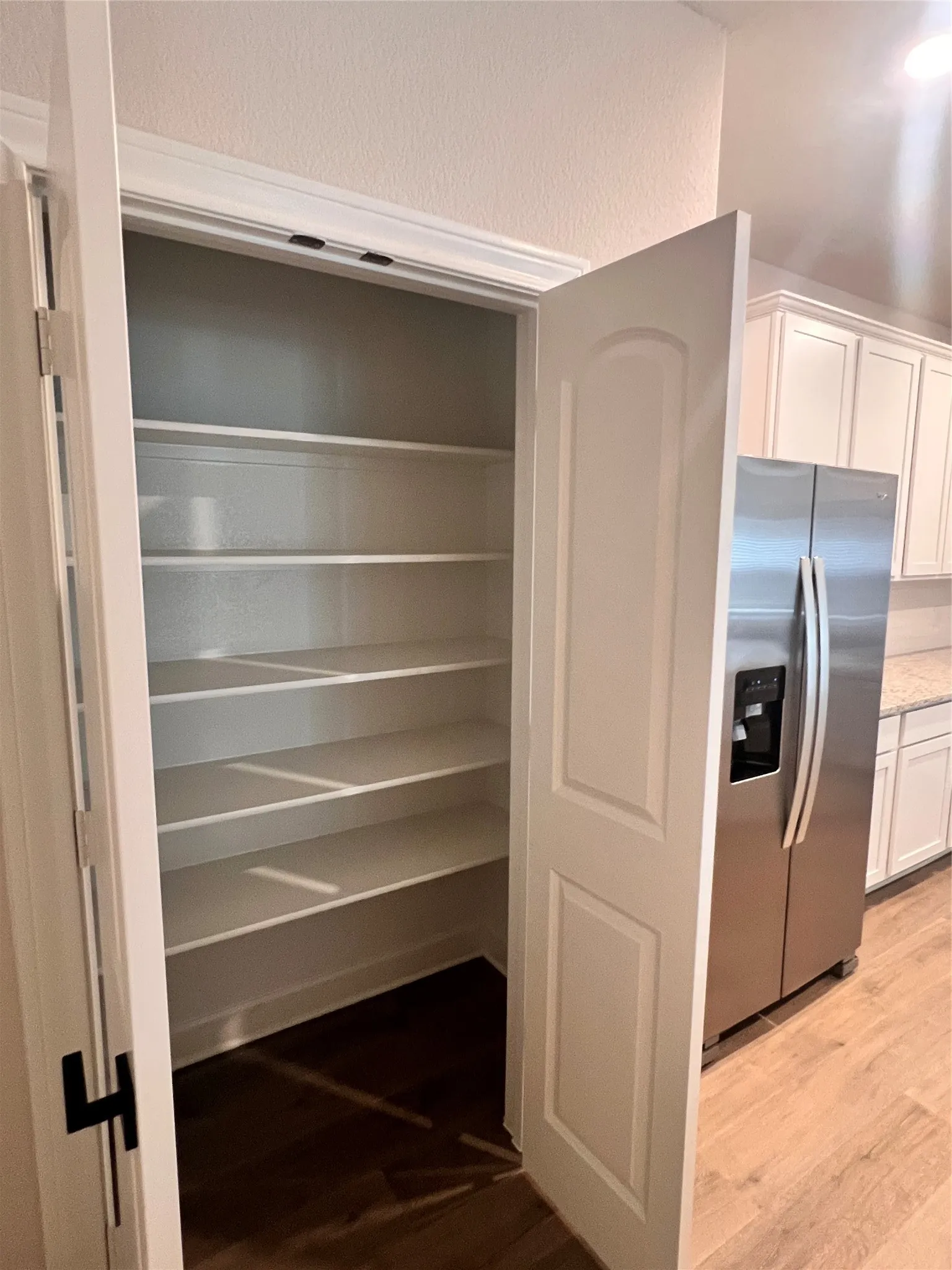Walk-in pantry with multiple shelves, adjacent to a kitchen featuring a stainless steel refrigerator, white cabinetry, and granite countertops