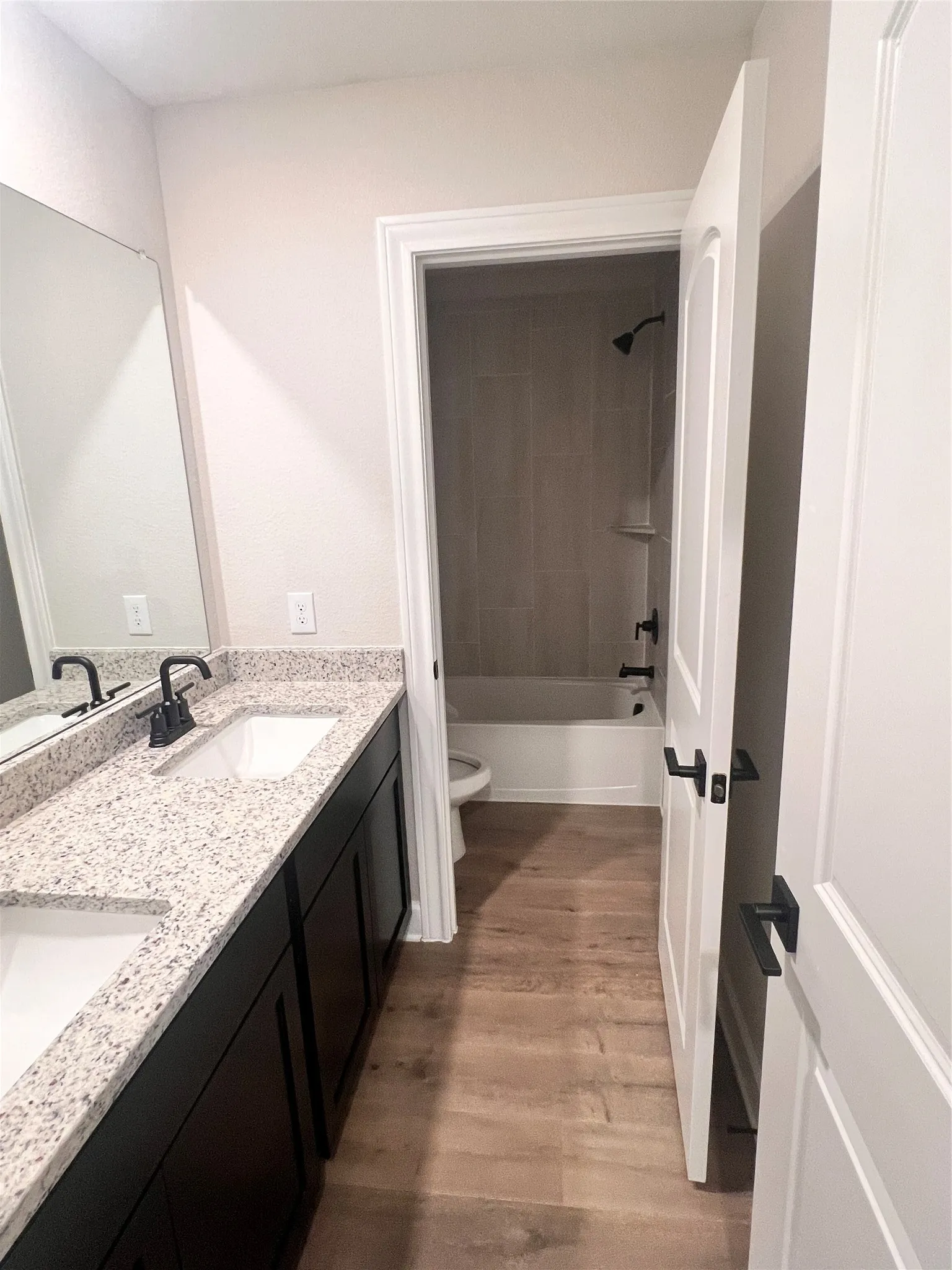 Primary bedroom with dual vanity with speckled countertop, wood-finish flooring, matte black fixtures, and a tiled shower/tub combination