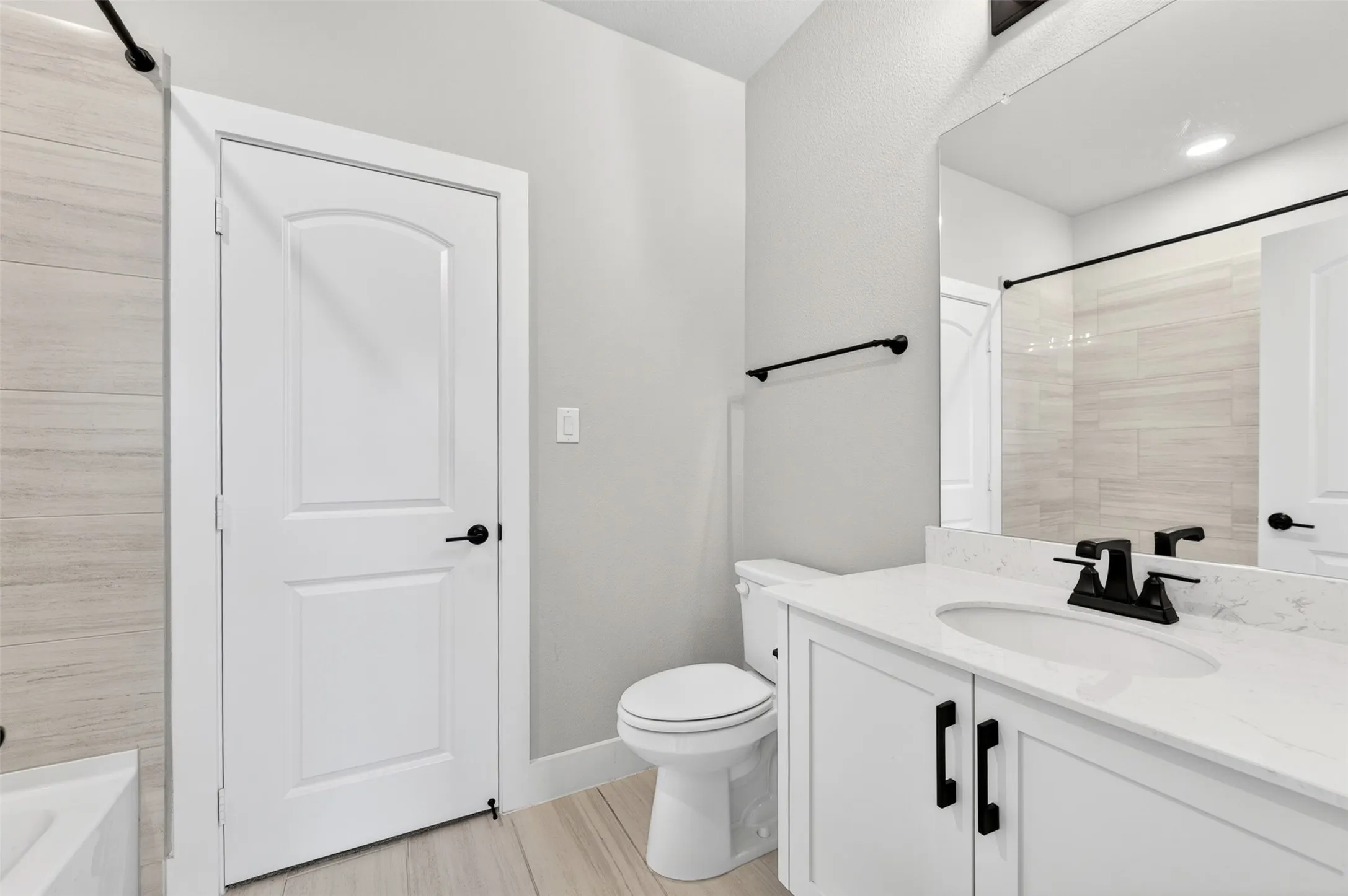 Light-toned plank flooring, white vanity with integrated sink, stone-finish countertop, matte black fixtures, and light-toned subway tile shower surround