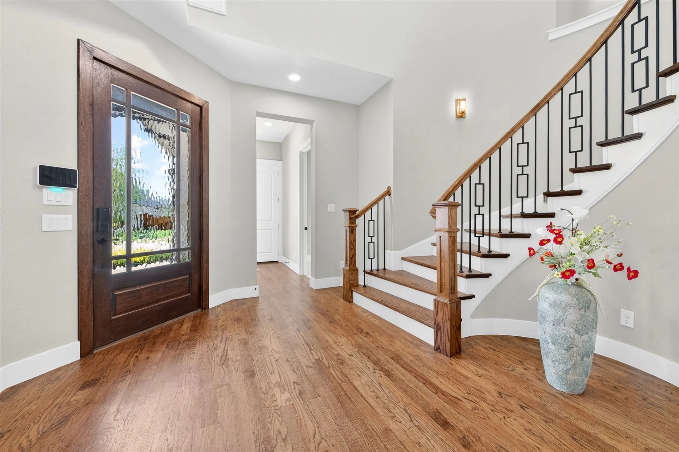 Wood-finish flooring, staircase with dark wood treads and white risers, decorative metal balusters with a wood handrail, wood-panel entry door with textured glass, and a wall sconce