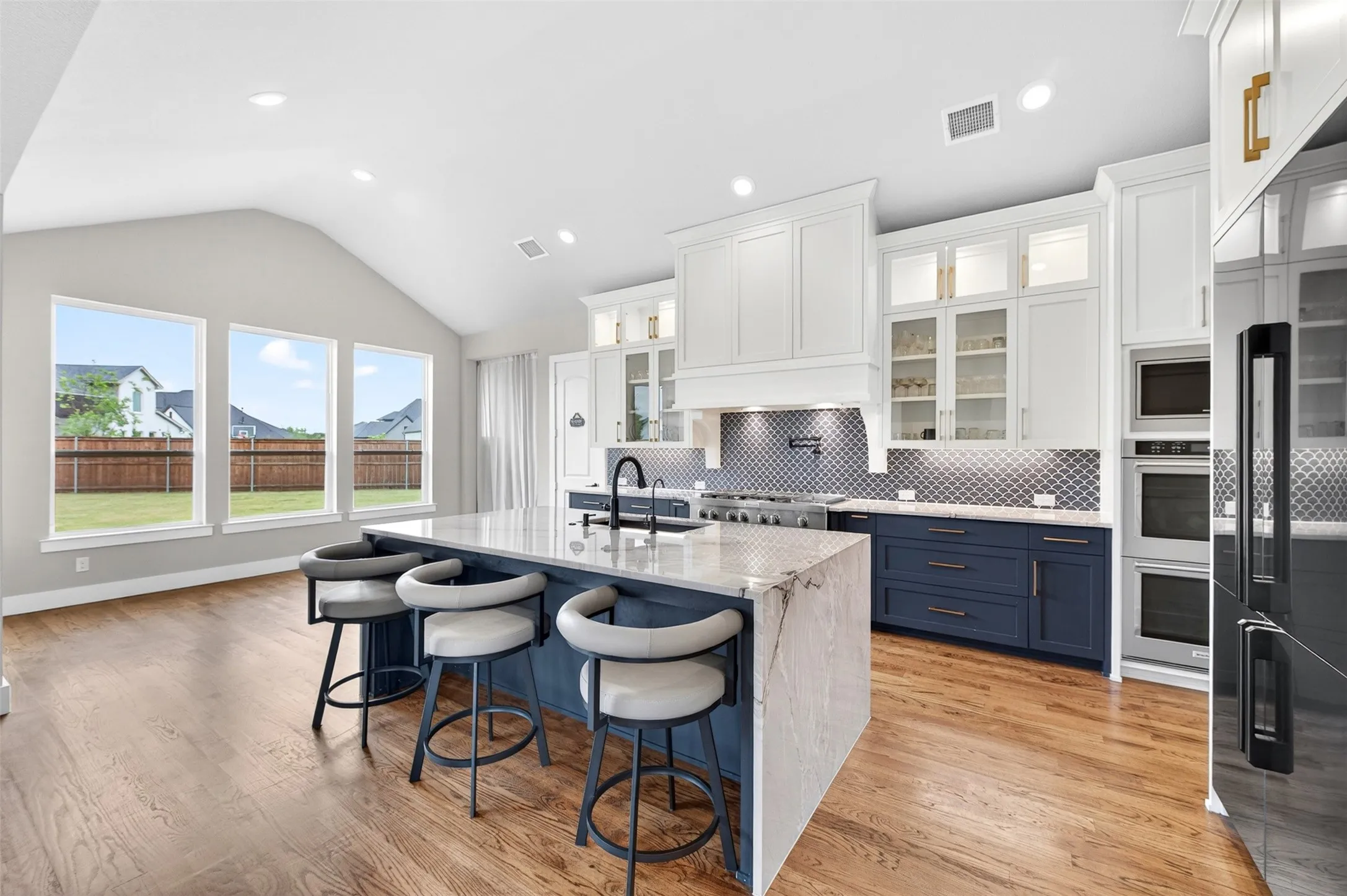 Vaulted ceiling over a kitchen featuring an oversized island with waterfall countertop, wood-finish flooring, two-tone cabinetry, and a full suite of stainless-steel appliances