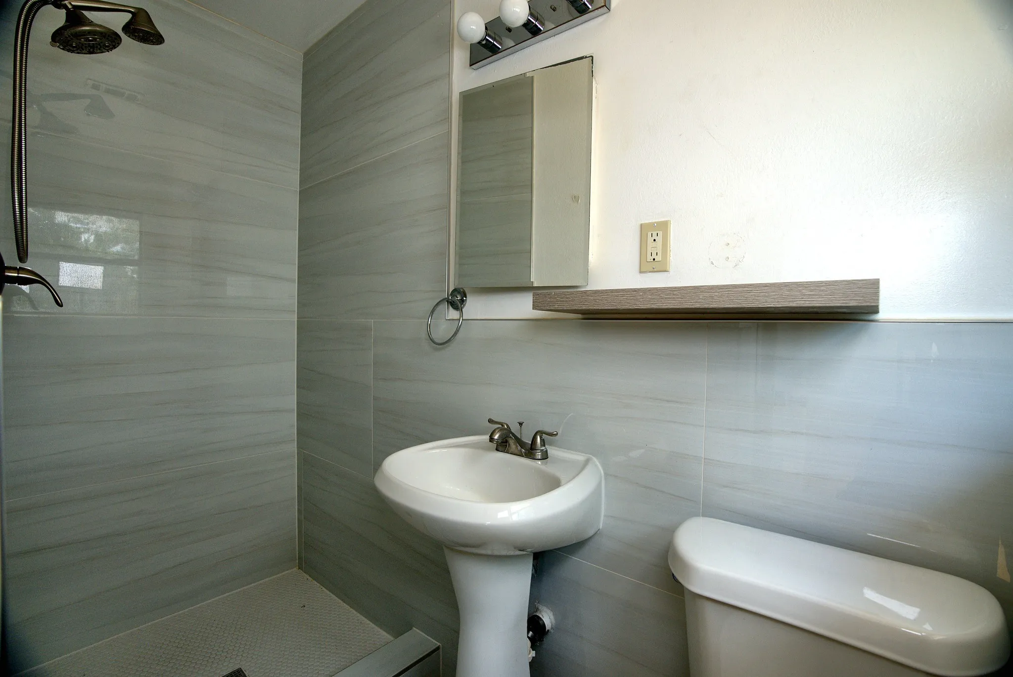 White pedestal sink with a dual-handle faucet, white toilet, and tiled shower surround featuring a bronze-finish shower head