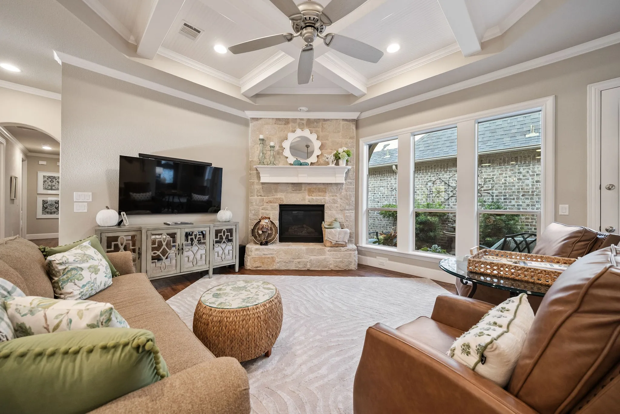 Coffered ceiling with a ceiling fan, stone-surround fireplace with a mantel, multiple large windows, and dark wood-finish flooring