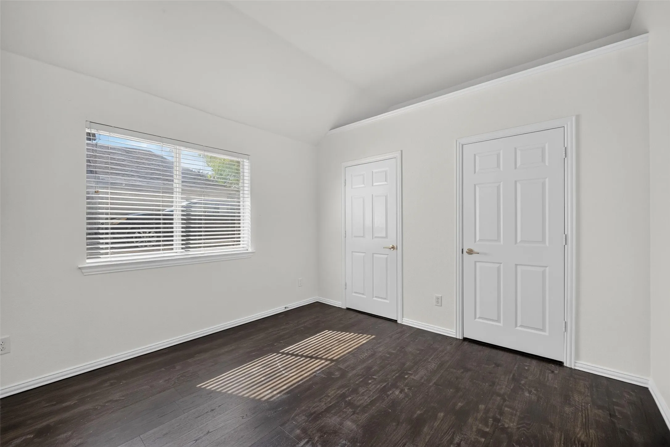Vaulted ceiling, dark wood-finish flooring, white paneled doors, crown molding, and a window with blinds