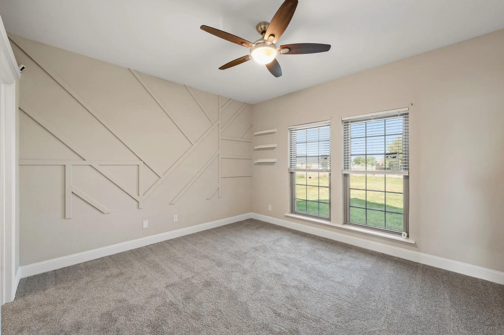 Geometric accent wall with decorative trim, two windows with blinds, gray carpet flooring, ceiling fan with integrated lighting, and three floating shelves