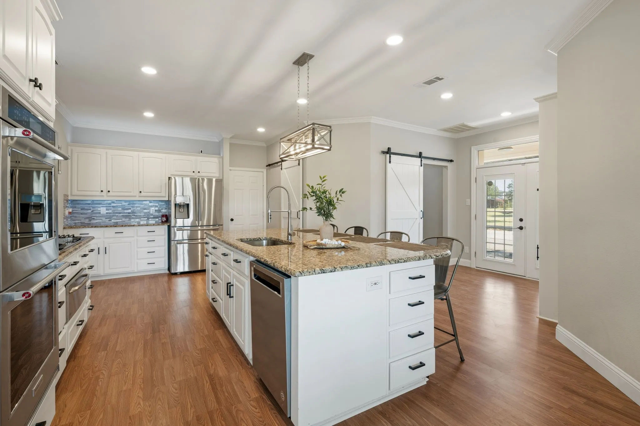 Spacious kitchen featuring an expansive center island with a sink, white cabinetry with dark hardware, stainless steel appliances, and wood-finish flooring