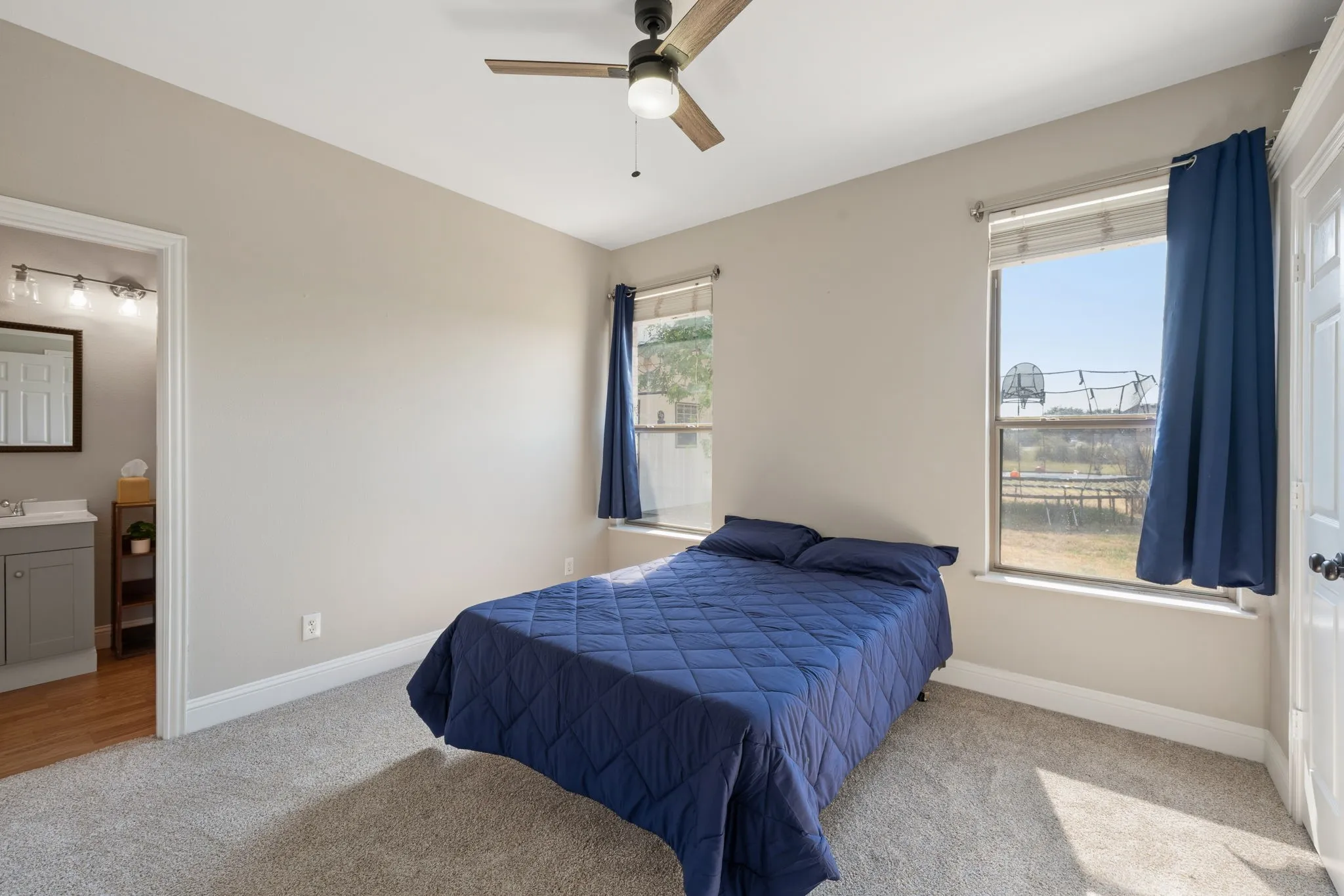 Carpeted flooring, two windows, ceiling fan with integrated lighting, and a doorway opening to a bathroom with a vanity and wood-finish flooring