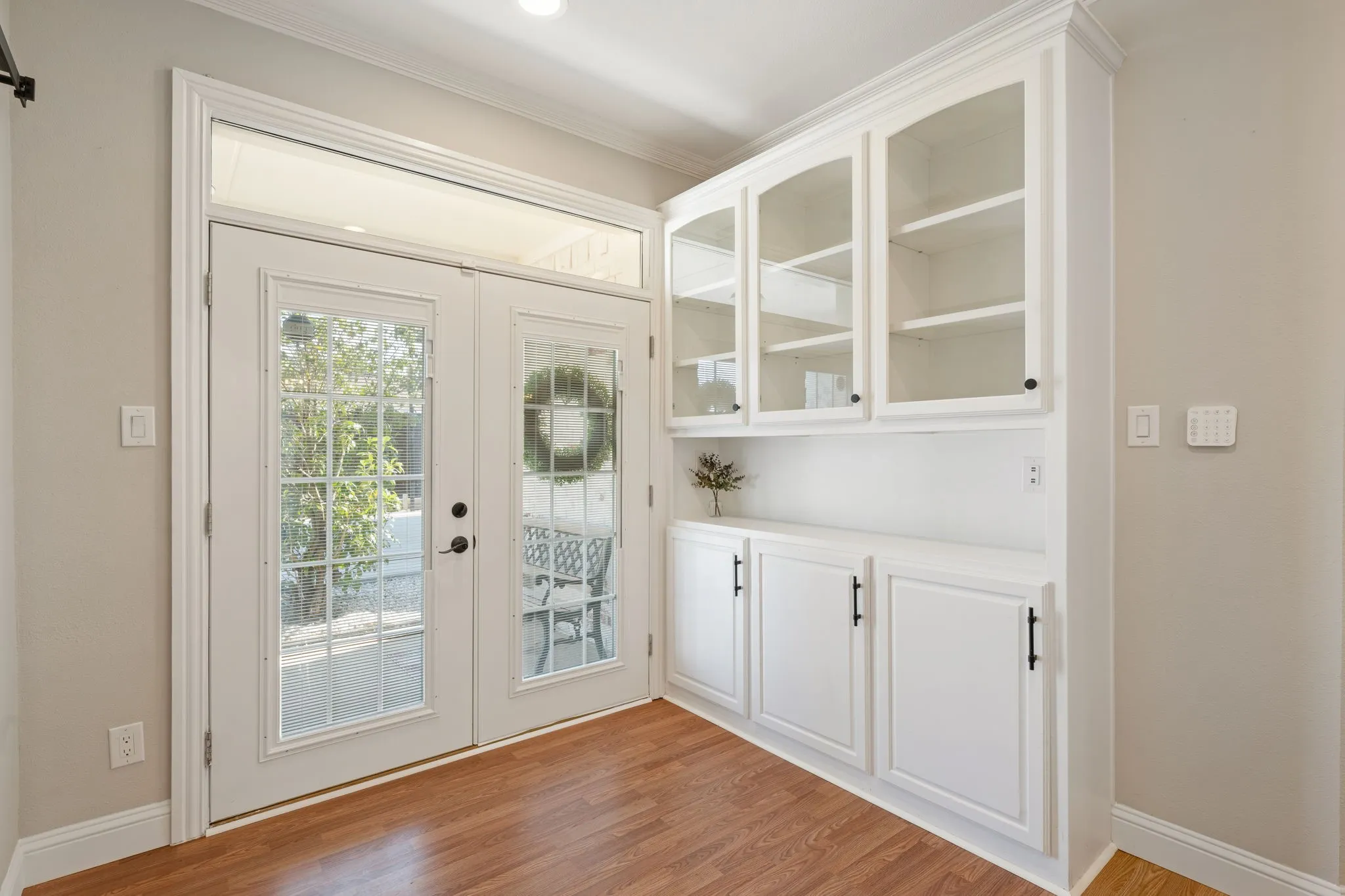 Double French doors with transom window, extensive built-in cabinetry with glass-front uppers, wood-finish flooring, and crown molding