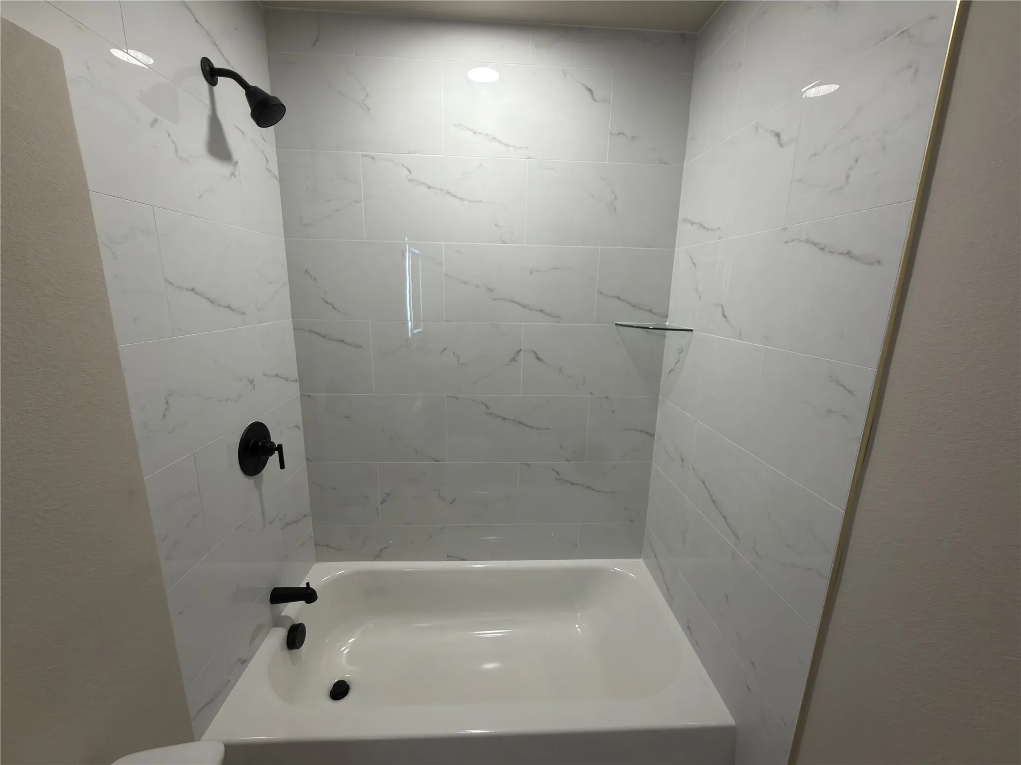 Bathtub with white tiled surround, matte black shower fixtures, and recessed glass shelf