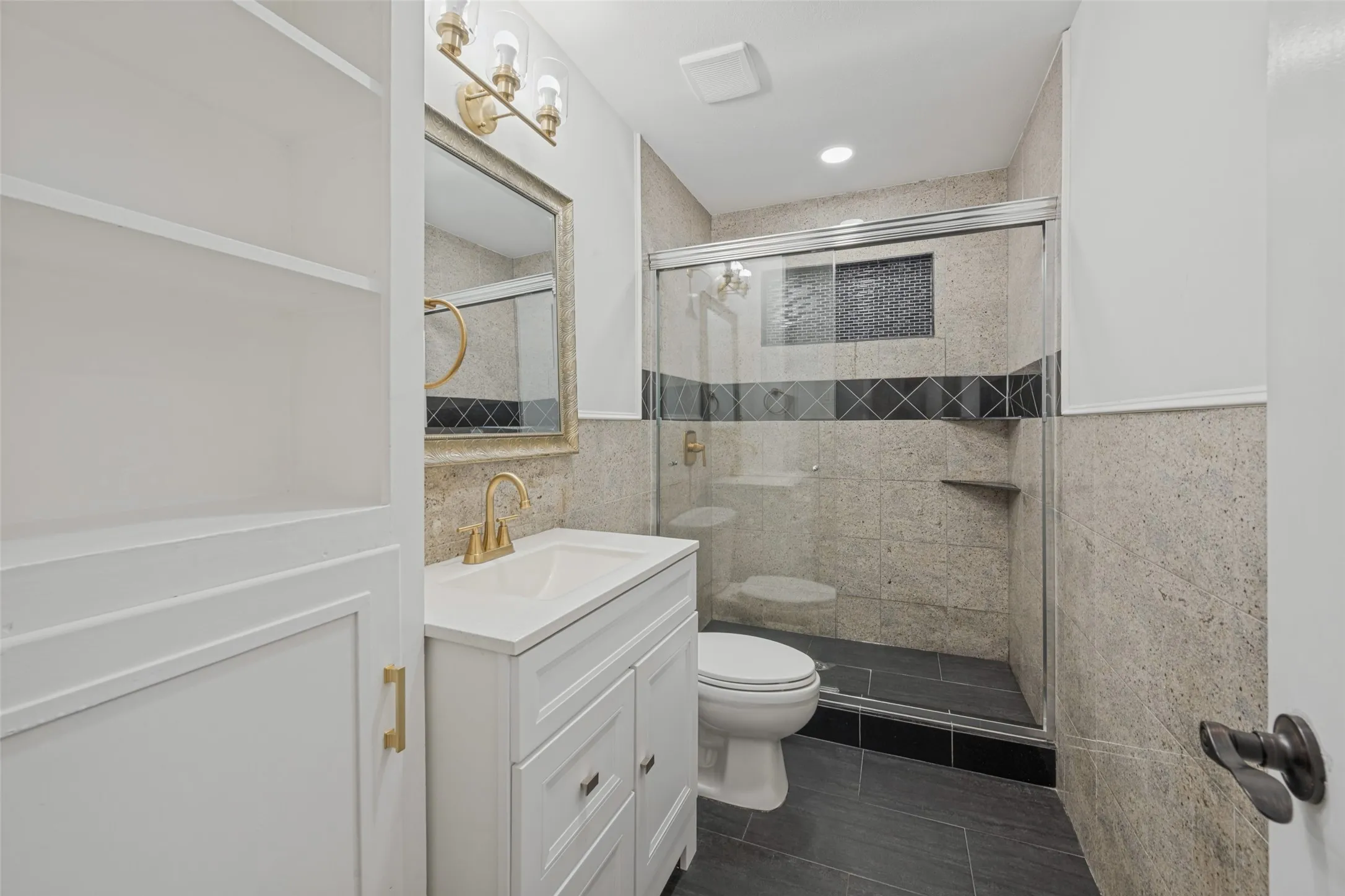Built-in shelving, vanity with white shaker-style cabinetry, gold-finish faucet, and a glass-enclosed shower with tiled walls