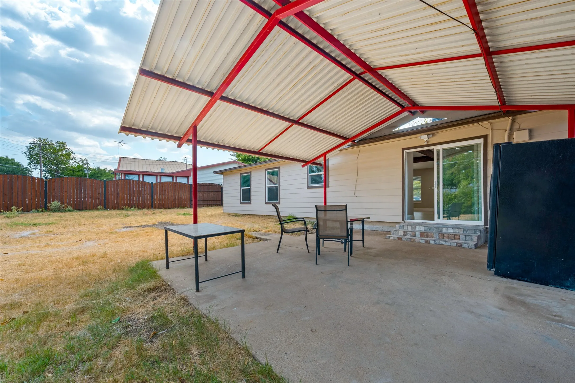 Concrete patio with an attached metal awning featuring red supports, leading to a sliding glass door entry with brick steps