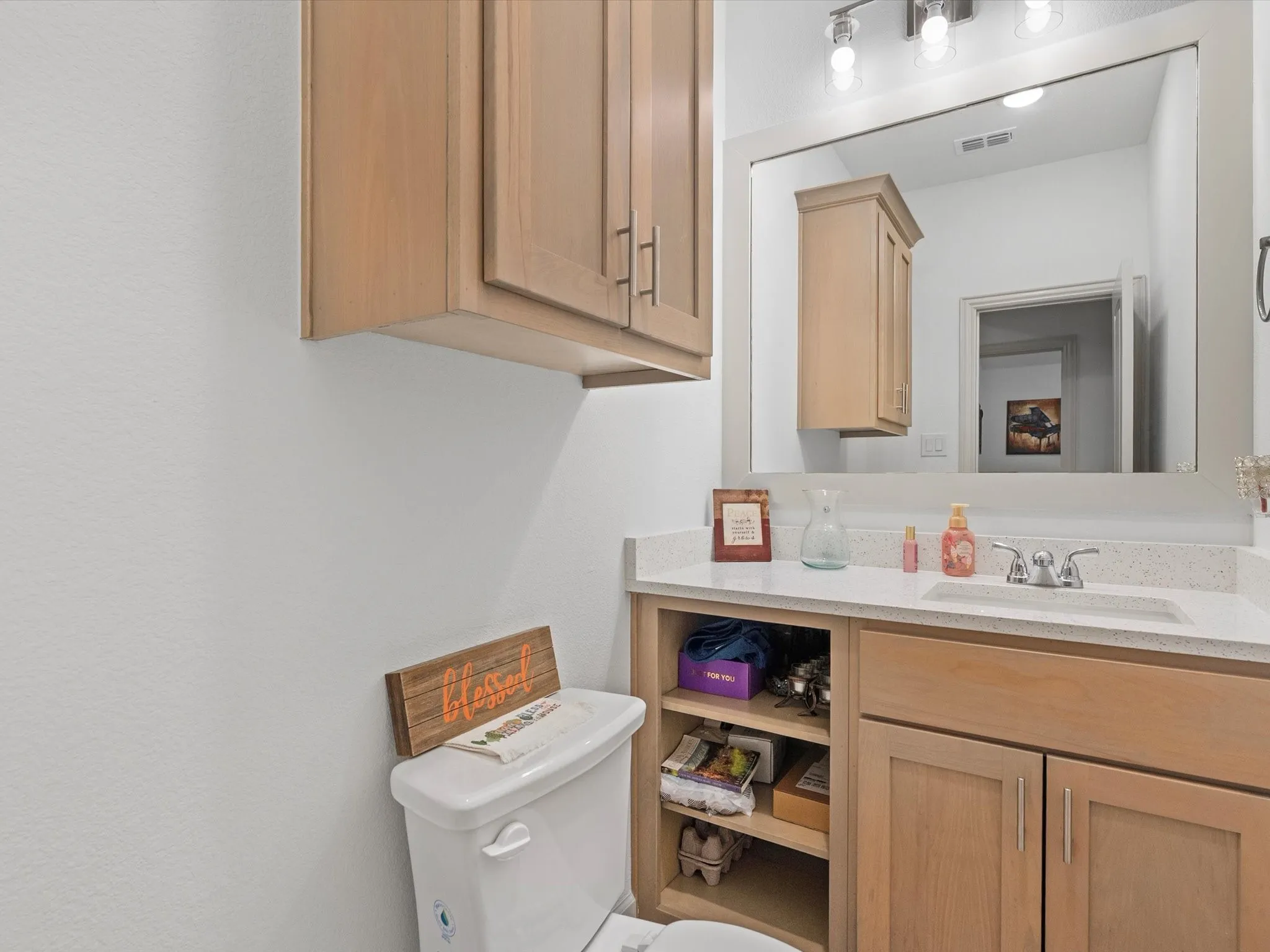 Wood-finish cabinetry with brushed nickel hardware, white countertop with integrated sink, chrome-finish faucet, and a wall-mounted mirror with overhead lighting