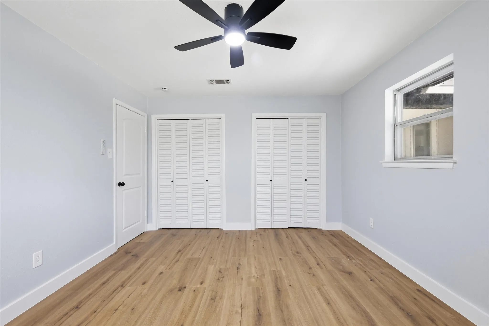 Room featuring wood-finish flooring, two sets of bi-fold louvered closet doors, a black ceiling fan with integrated lighting, and a white trim window