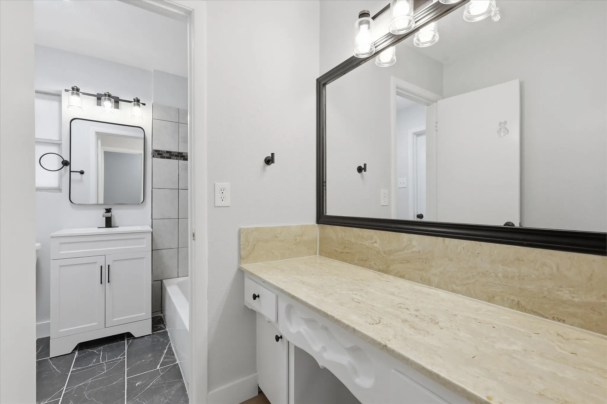 Dual bathroom vanity spaces featuring a light-toned countertop, a large framed mirror with overhead lighting, white cabinetry, and matte black hardware