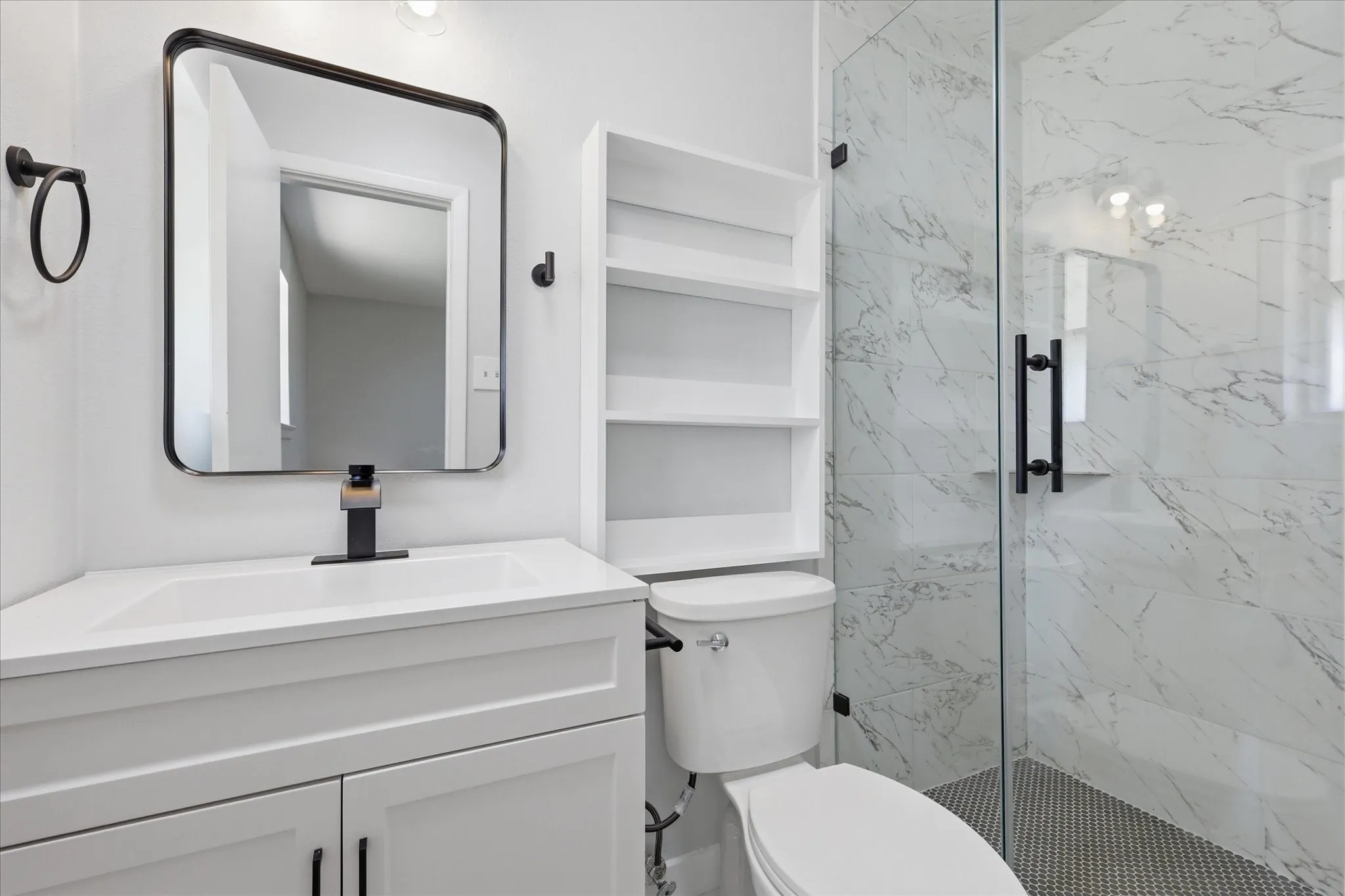 Modern bathroom featuring a white vanity with integrated sink, matte black fixtures, a frameless glass shower enclosure with marble-look wall tiles, and hexagon mosaic floor tiles