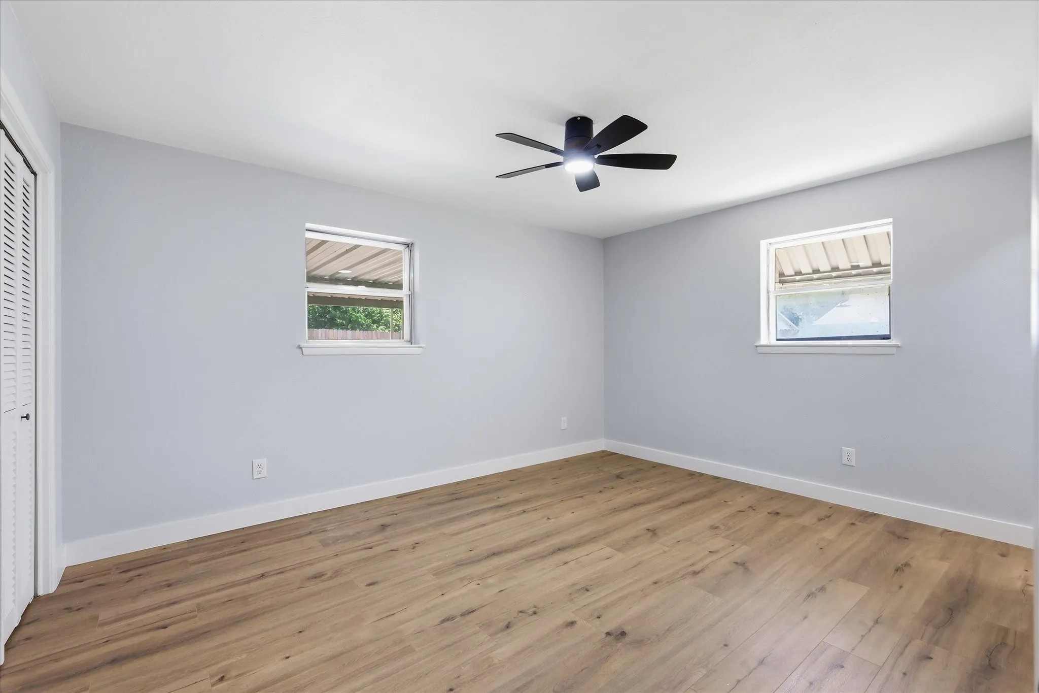 Spacious room featuring light wood-finish flooring, a modern black ceiling fan, and two windows providing natural illumination
