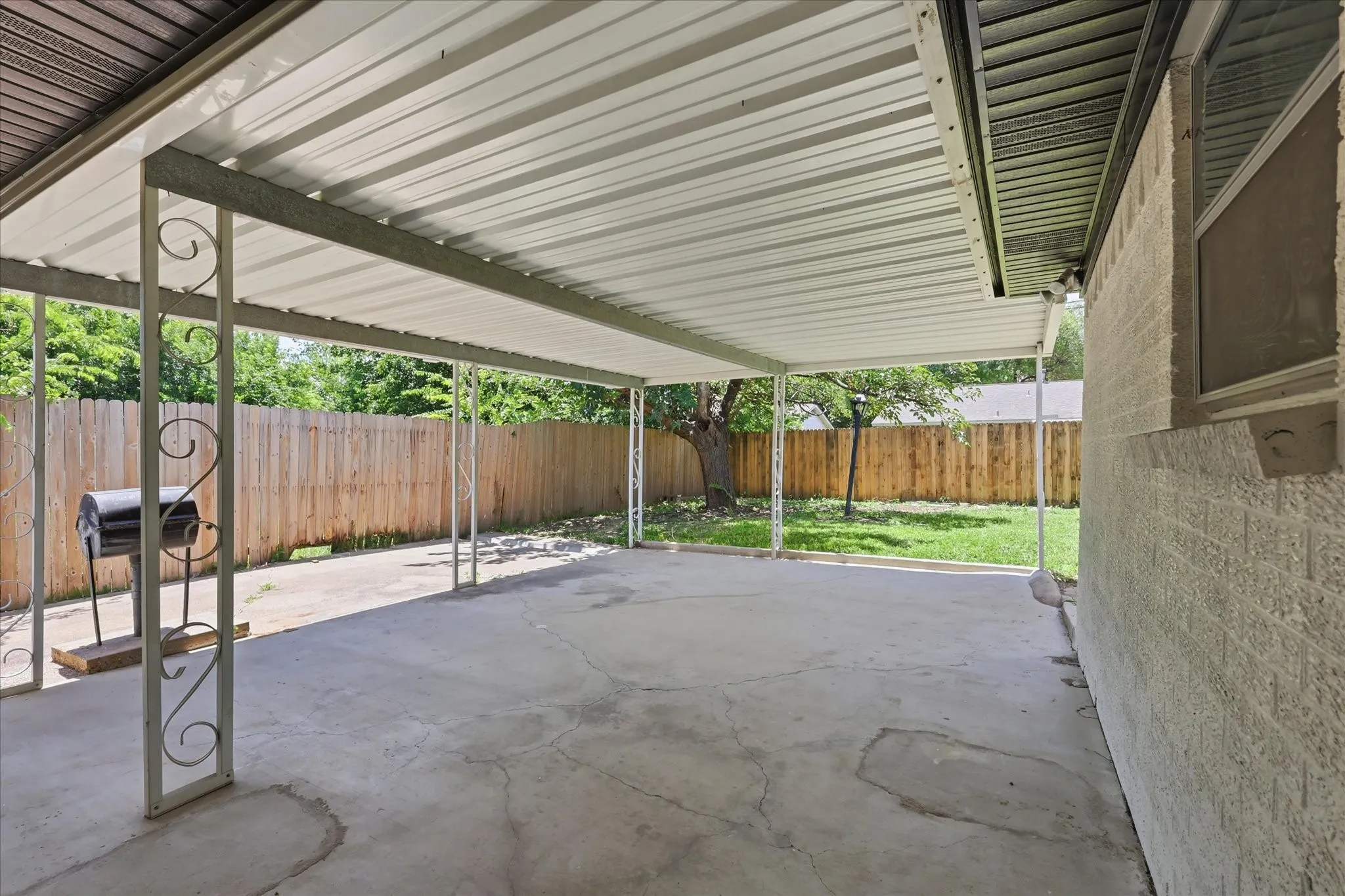 Expansive covered patio featuring a corrugated metal roof, decorative metal support columns, a concrete slab, and a wood privacy fence